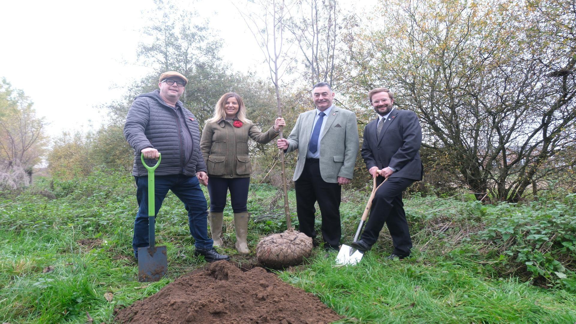Three men and a woman stand on a patch of green grass behind a mound of brown earth. In the centre, a woman with blond hair, dressed in a green jacket, dark trousers and grey wellies, holds a bare tree sapling with a man standing next to her, who wears a grey blazer, black trousers, a pale shirt and a blue tie. To the left, a man wearing a flat cap, blue coat and blue jeans holds a spade with a bright green handle. To the right, a man with short brown hair and a beard, dressed in a dark suit, poses with his foot on a spade as if preparing to dig.