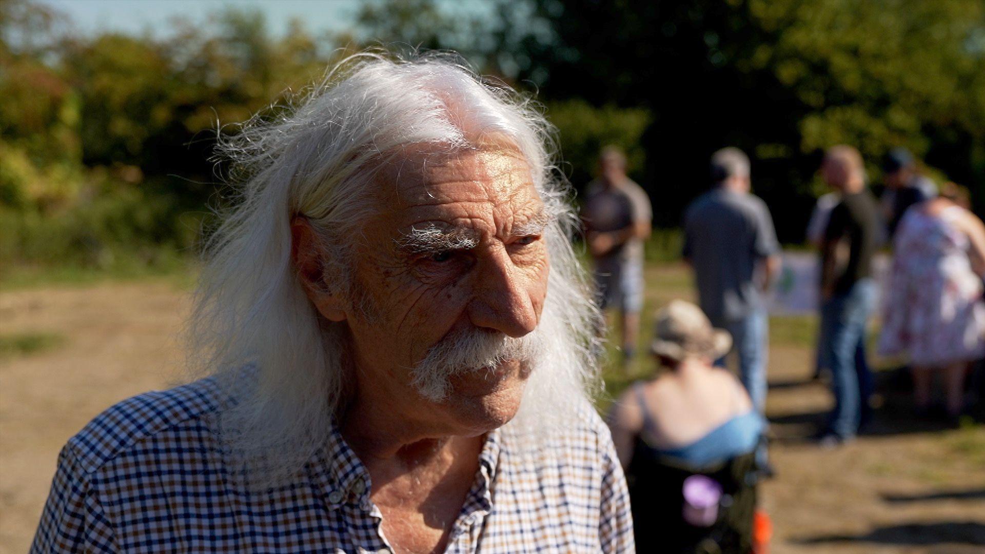David Freeman is an older man with shoulder-length white hair and a moustache. He is wearing a blue checked shirt. People can be seen blurred in the background