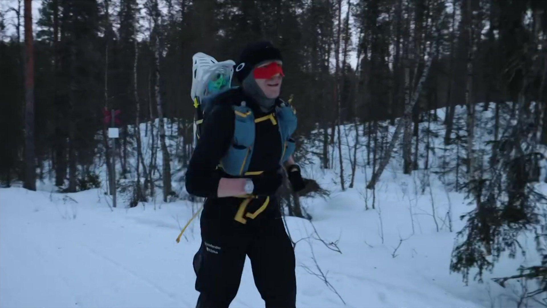 Alex is surrounded by trees and runs through thick snow. He wears all black, red goggles and a blue rucksack with yellow straps