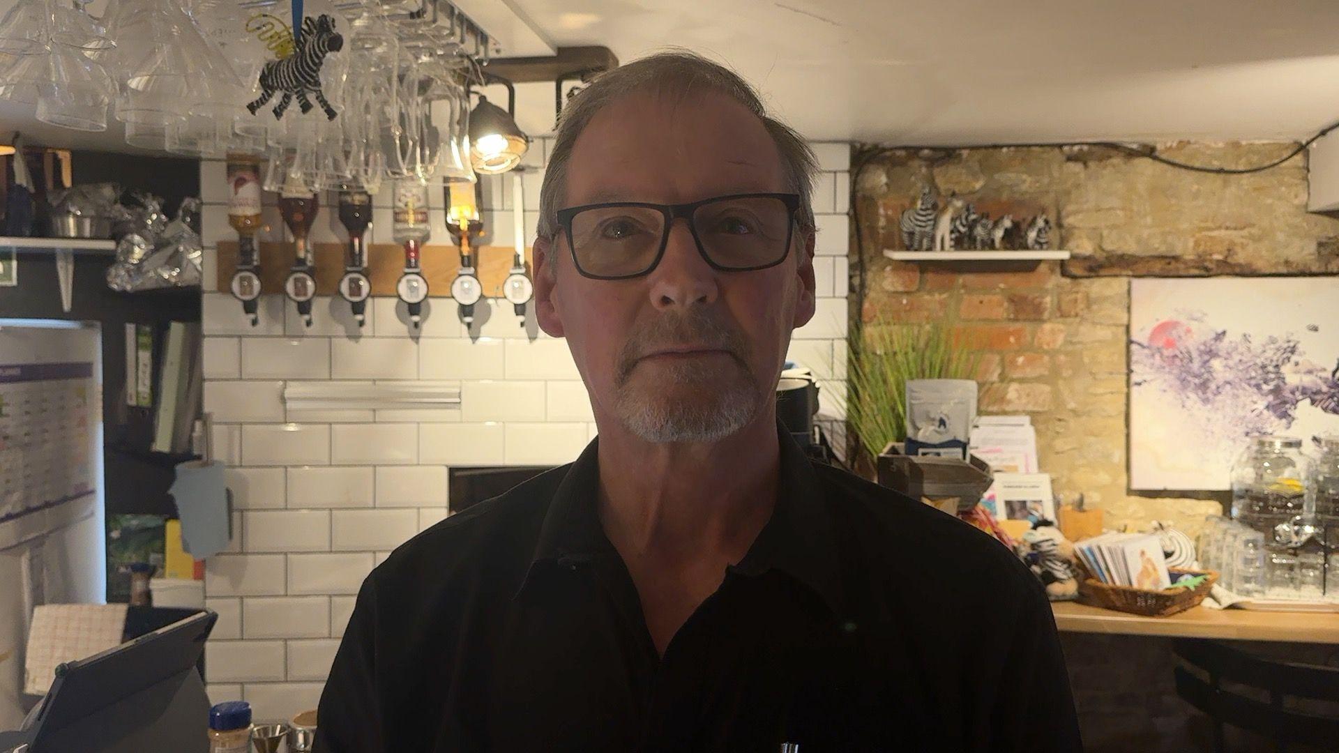 Johnny Morgan is looking at the camera and is stood inside his cafe. He has glasses and is wearing a black polo shirt.
