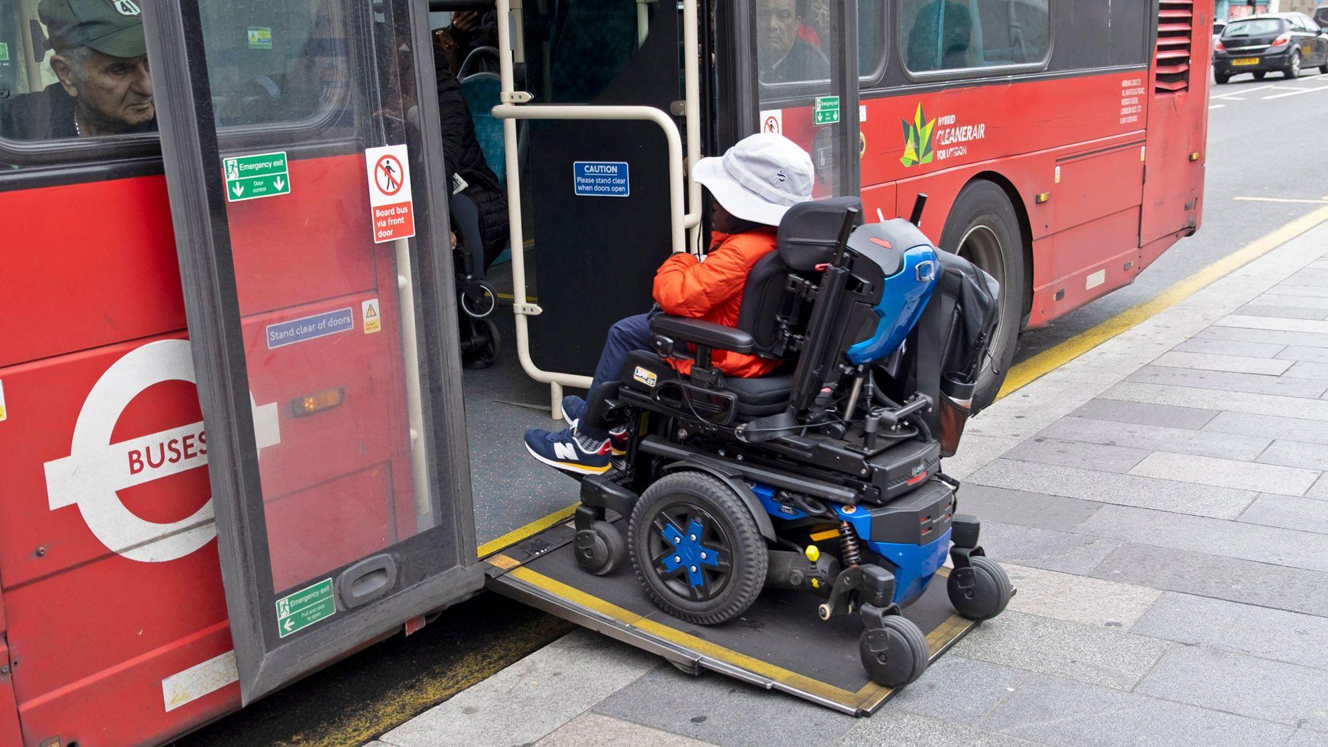 A person in a motorised wheelchair wearing an orange jacket and a white hat boards a bus in London using a wheelchair ramp in October 2025