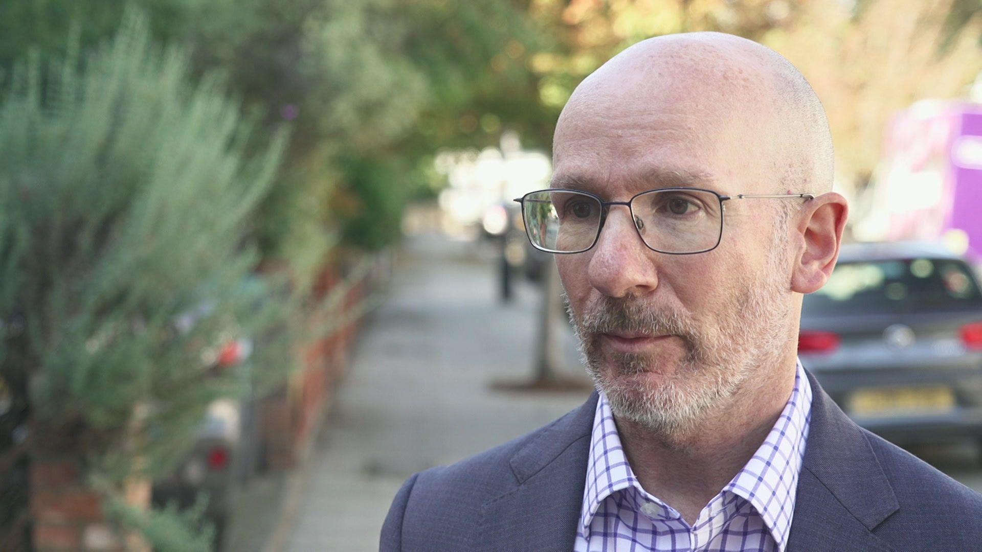 Man looking towards the the left, wearing dark metal rimmed glasses, he has a light white and brown beard, is bald and wears a light blue/grey suit and a white and purple checked shirt. He's standing on a pavement and to the left is a low brick wall, with green bushes overhanging the brick wall.