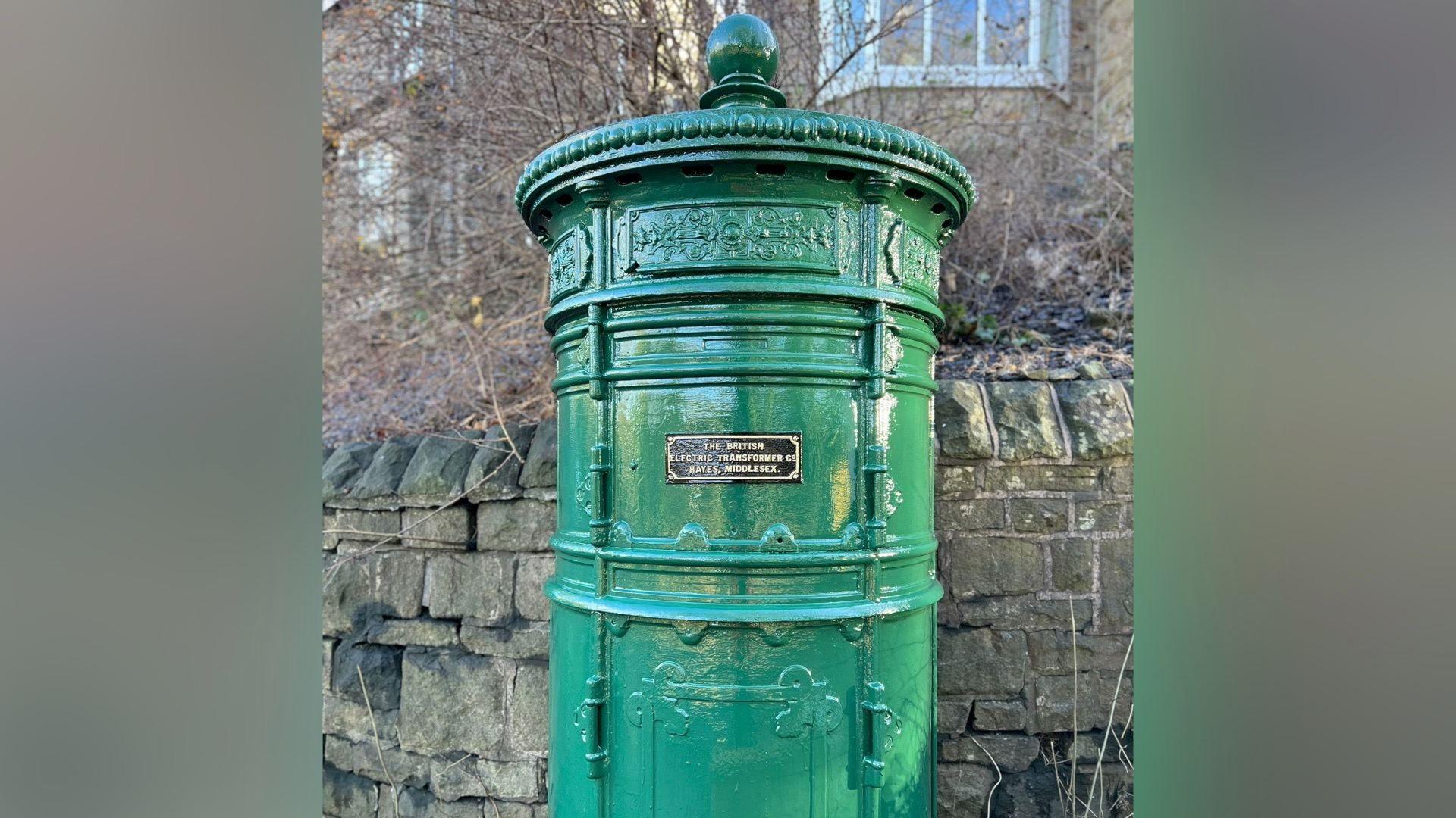 A freshly-painted bright green electricity distribution pillar with a black plaque fastened to its centre which reads 'The British Electric Transformer Co, Hayes, Middlesex'. It stands before a sandstone weathered wall with houses and empty branches in the background.