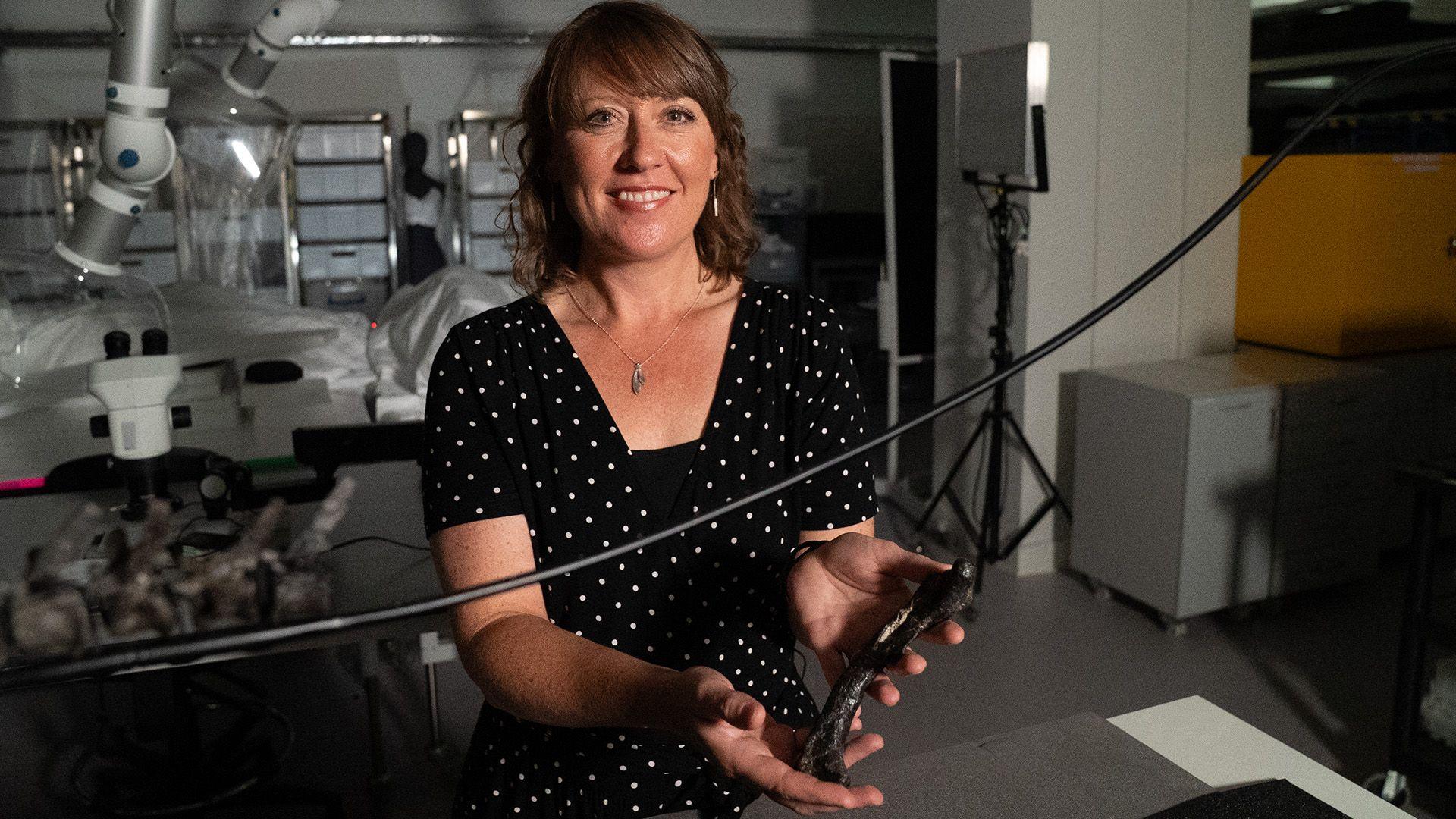 A woman with brown hair looking at the camera, holding what appears to be a dinosaur bone. She is smiling, standing in a science lab, wearing a navy dress with small white spots