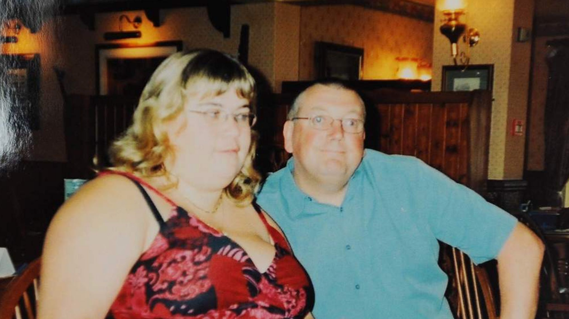 Allison and Phil Heathcote are photographed sitting down. Allison, who is looking away from the camera, is wearing a red patterned dress and Phil, who is smiling at the camera, is in a blue shirt.