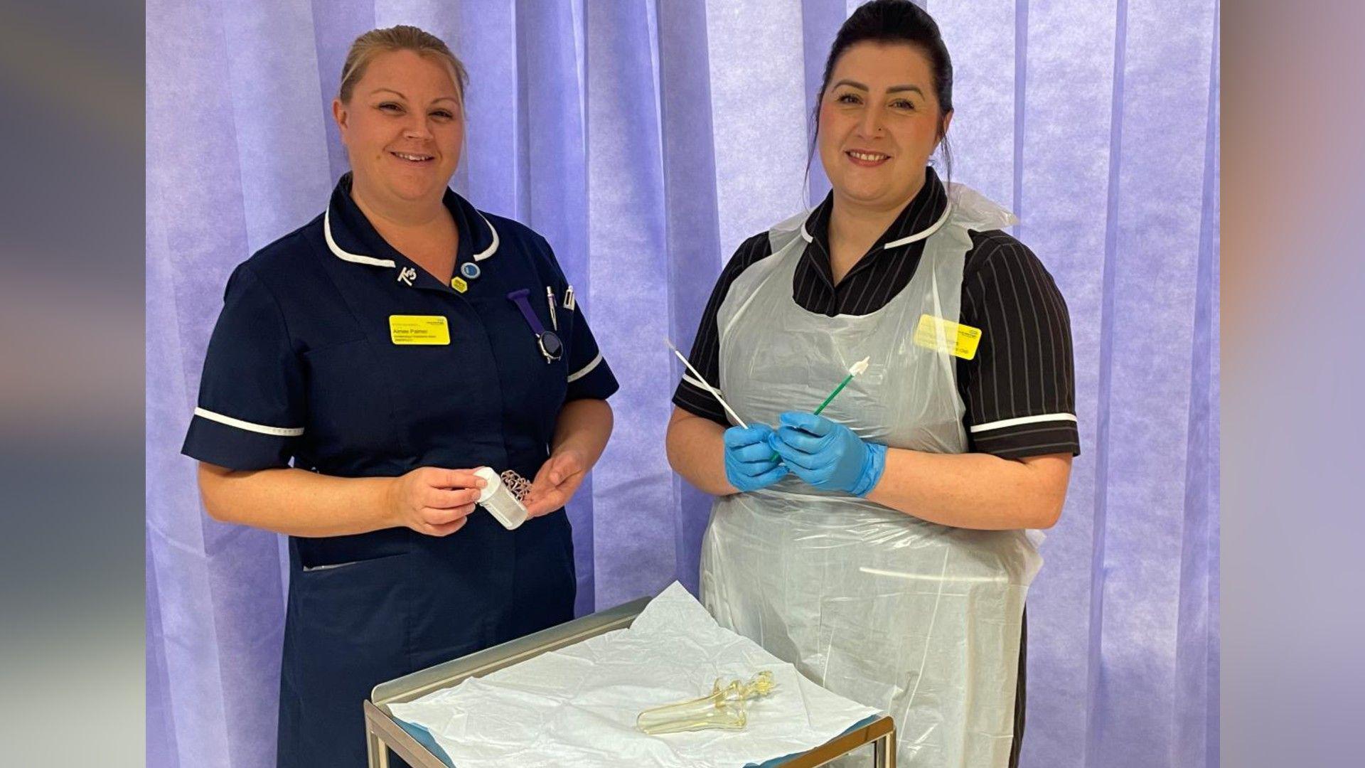 Two nurses stand before a purple hospital curtain. They are both wearing nurses uniform and are holding up instruments used during the cervical smear test. In front of them is a silver tray with another piece of equipment. They are both smiling at the camera. 