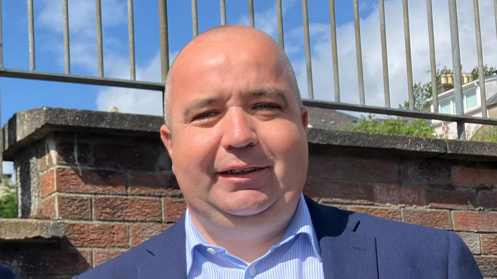 Brian Tierney is seen in a light blue and whiteshirt and navy blazer. 
He is standing in front of a red brick wall on top of which is metal railings.