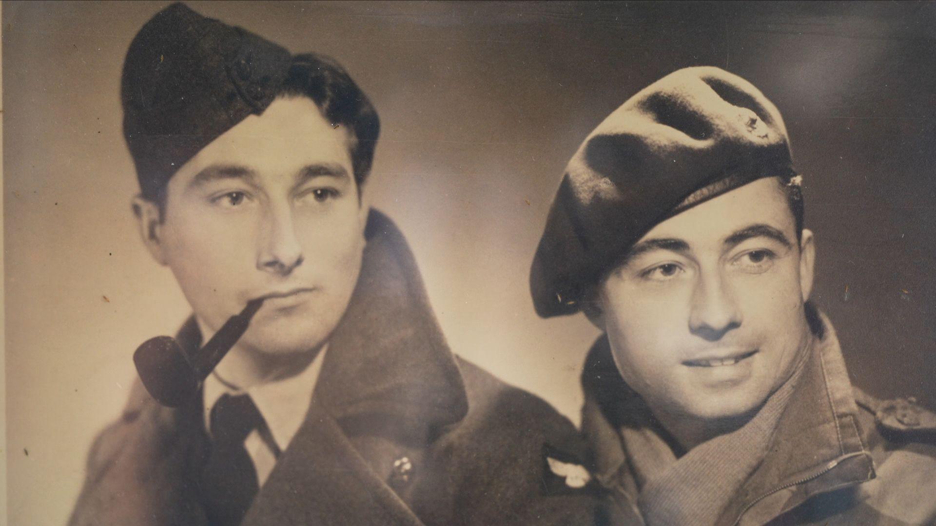 Black and white photo showing a man in air force uniform wearing a hat and smoking a pipe, next to a man in Royal Marines Commando uniform wearing a beret and scarf.