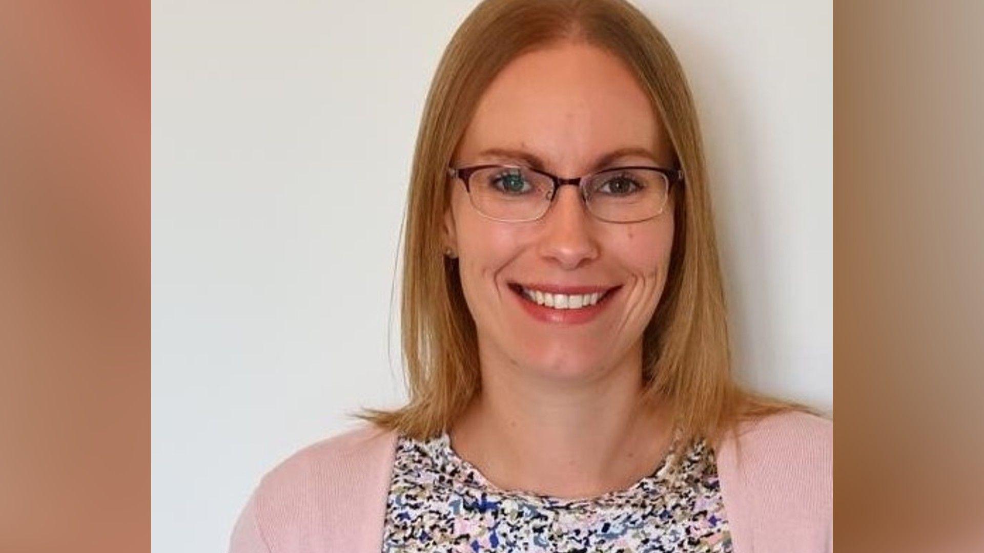 A head-and-shoulders picture of Dr Zoe Rawlings. She is smiling, has shoulder-length blond hair, glasses, and is wearing a pink cardigan and a colourful blouse. She has been photographed against a plain background.