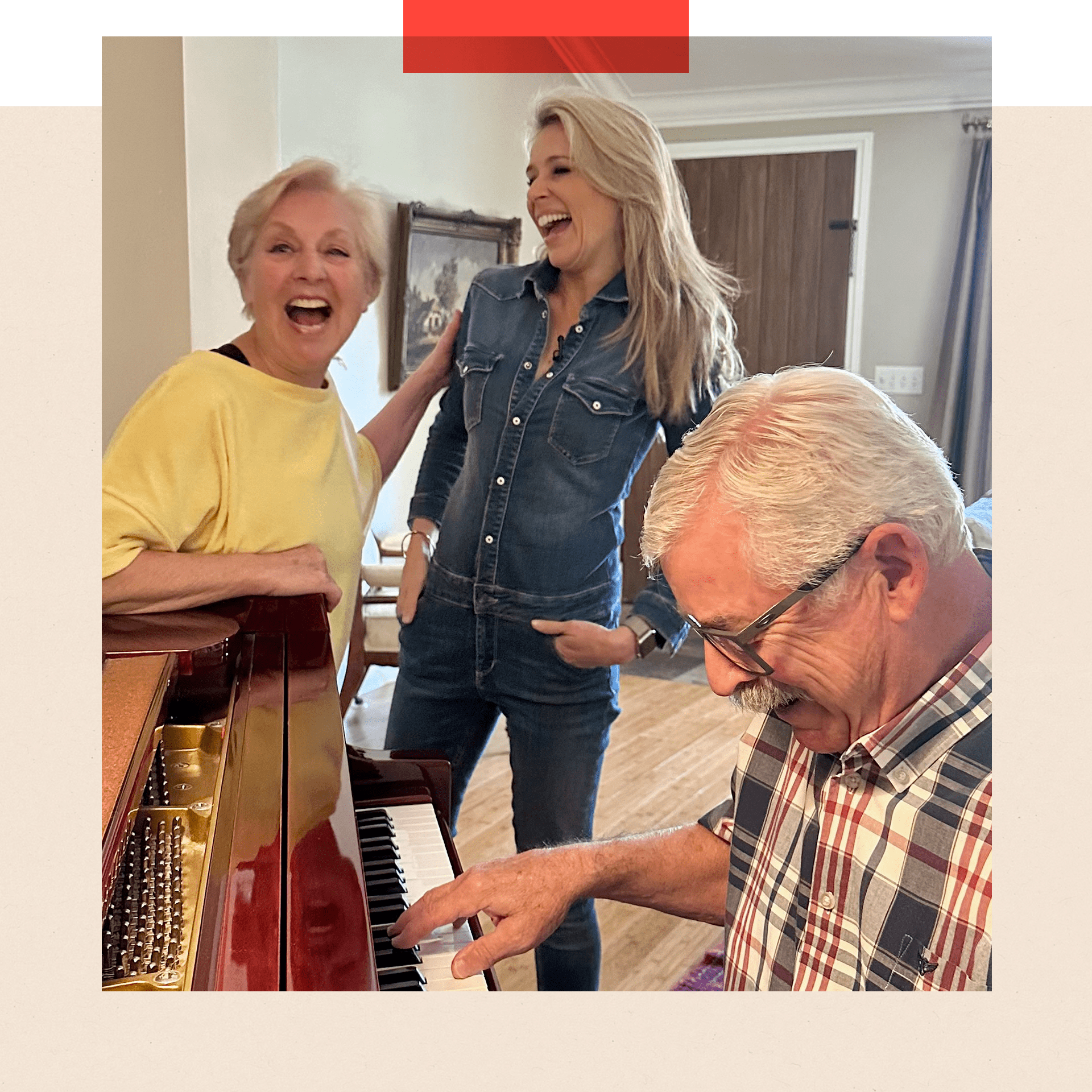 A smiling Marijke stands next to Lara Lewington while Marijke's husband Tom plays the piano