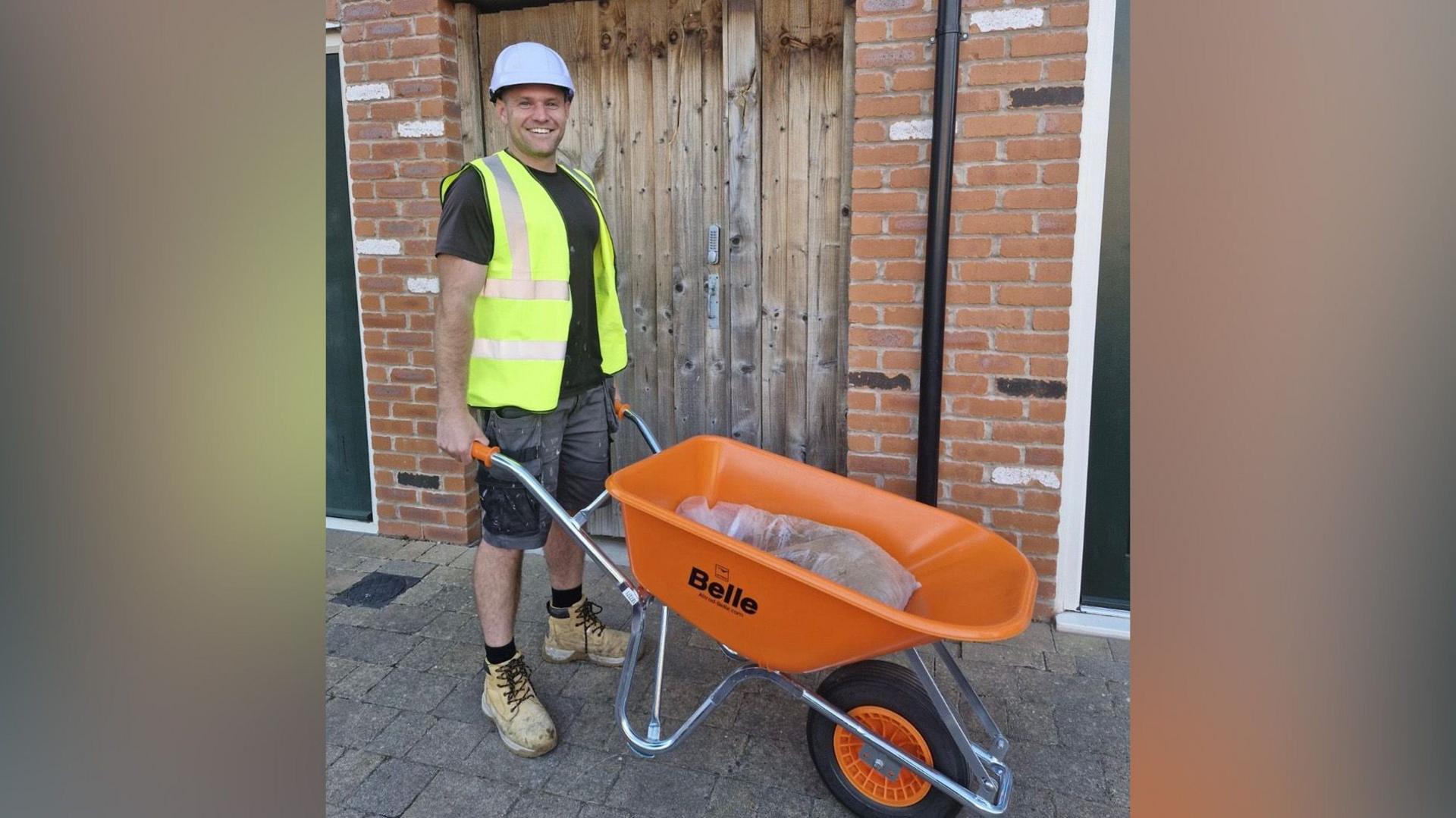 A man wearing a white helmet and hi-vis jacket over a black t-shirt, stands and smiles while standing in front of a wooden gate.