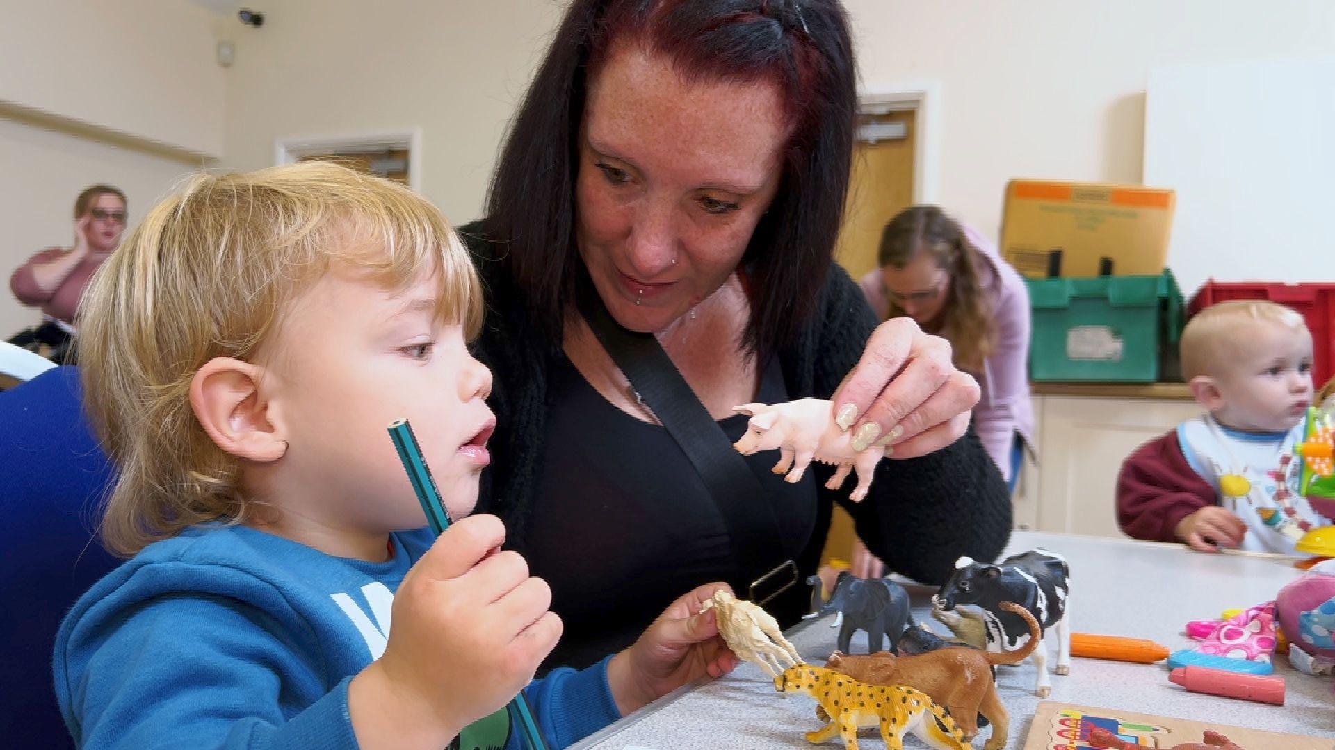 A toddler and his mum playing with toy animals at a table. Other families are in the background.