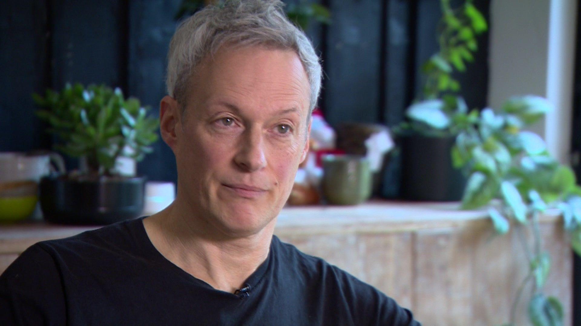 Andy Leary-May, wearing a black t-shirt, is seen in his house. A table with plants on it is pictured in the background.