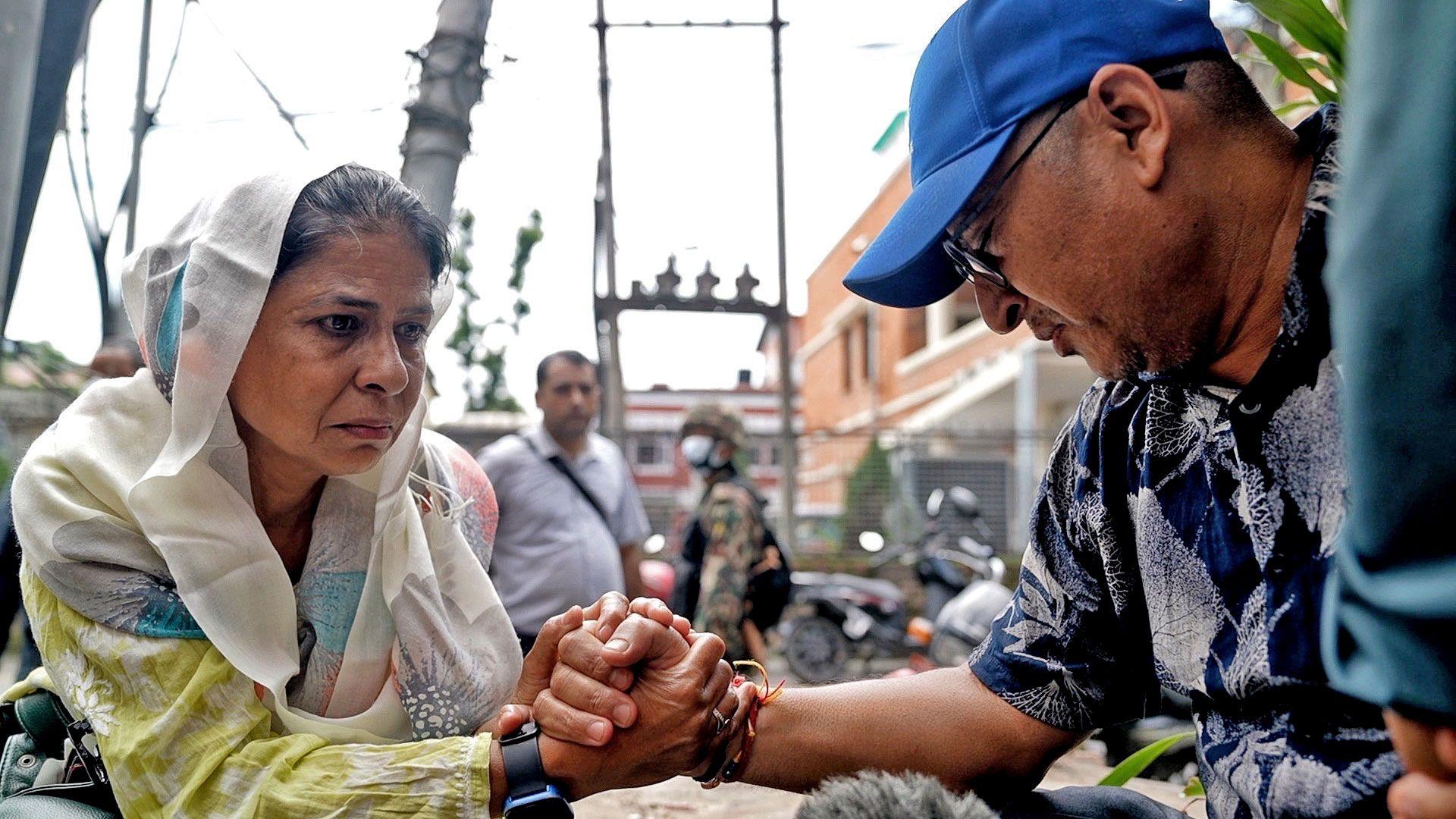 A man and a woman grasp each others hands, the man wears a blue cap, glasses and a short sleeved navy and white top. The woman wears a white and pale blue scarf on her head, and a yellow and white dress. Behind them a building and a soldier can be seen