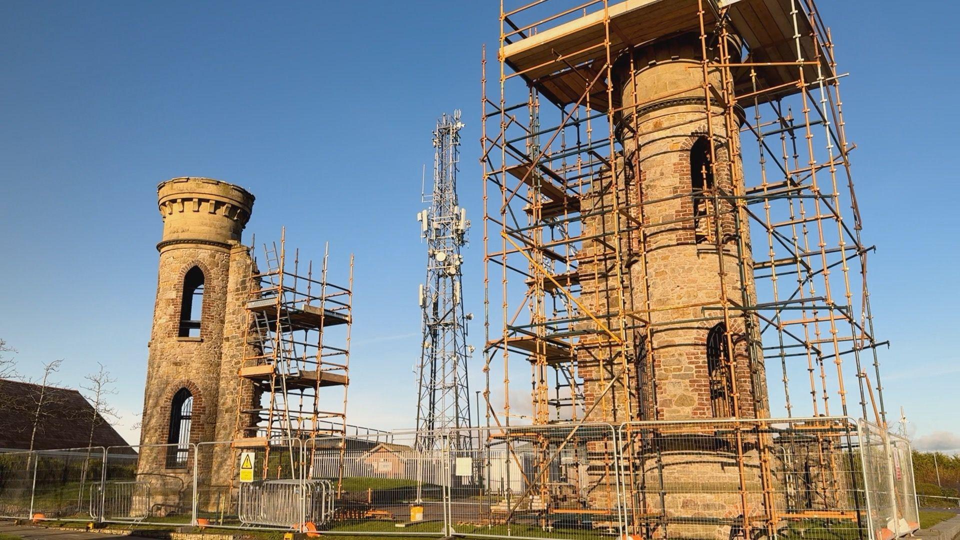 Two large stone pillars stand a distance apart in a green area. They are both surrounded by scaffolding and metal fences. In the middle is a large metal pylon.