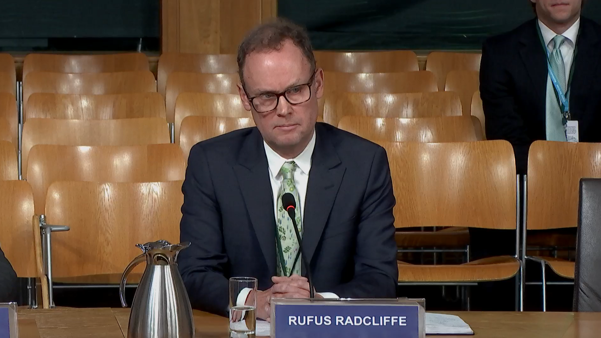 Rufus Radcliffe, who has thinning brown hair and black-rimmed glasses, sits with his hands clasped on a wooden table. He is wearing a dark suit, white shirt and green tie. He is sitting behind a placard with his name on it. There are rows of empty wooden seats in the background, with one man in a suit sitting behind him.