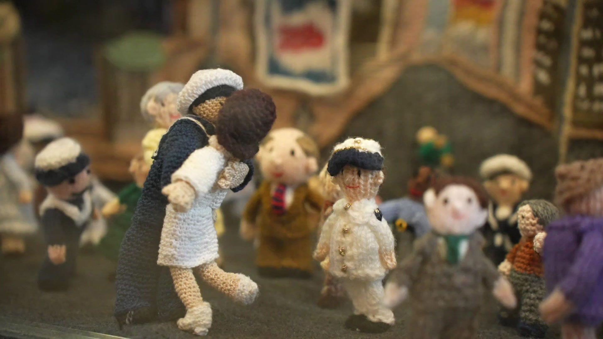 A depiction of the famous photograph of a navy sailor kissing a stranger in a white nurses uniform, made out of yarn. Several yarn people are in the background