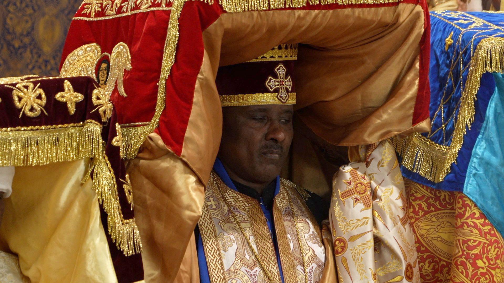 A man in traditional gold and blue robes, with material going over his head. He wears a round hat with a cross at the front.