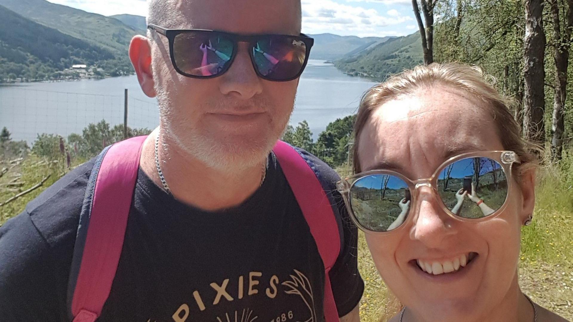 Leigh taking a selfie with her husband Colin on a sunny day near a lake while they are on holiday.