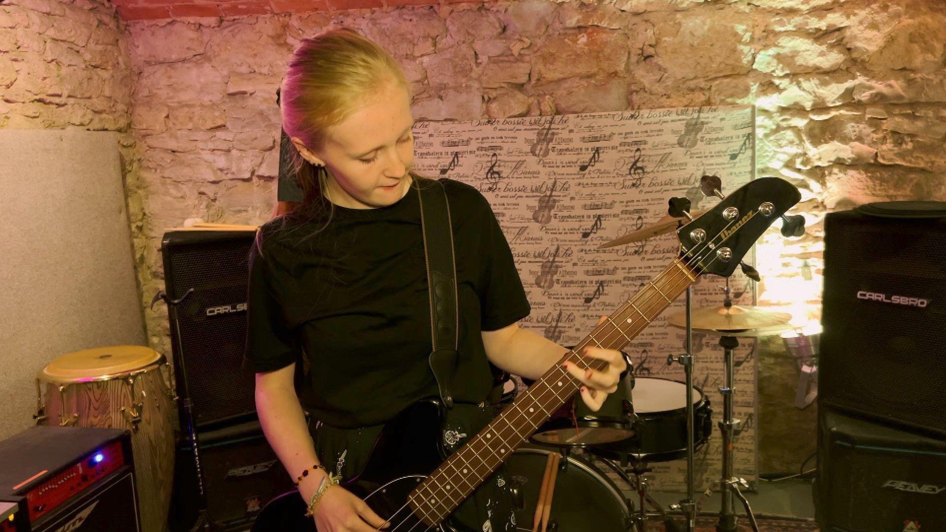 Teenage girl Ruby plays bass in the church crypt. There are white exposed brick walls in the background and a drum kit and amplifiers behind her.