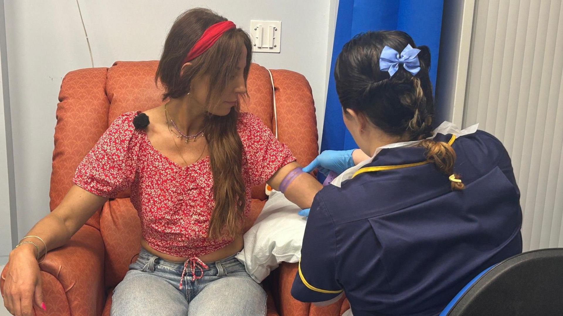 A woman sat on a hospital chair having her blood taken from her arm by a nurse