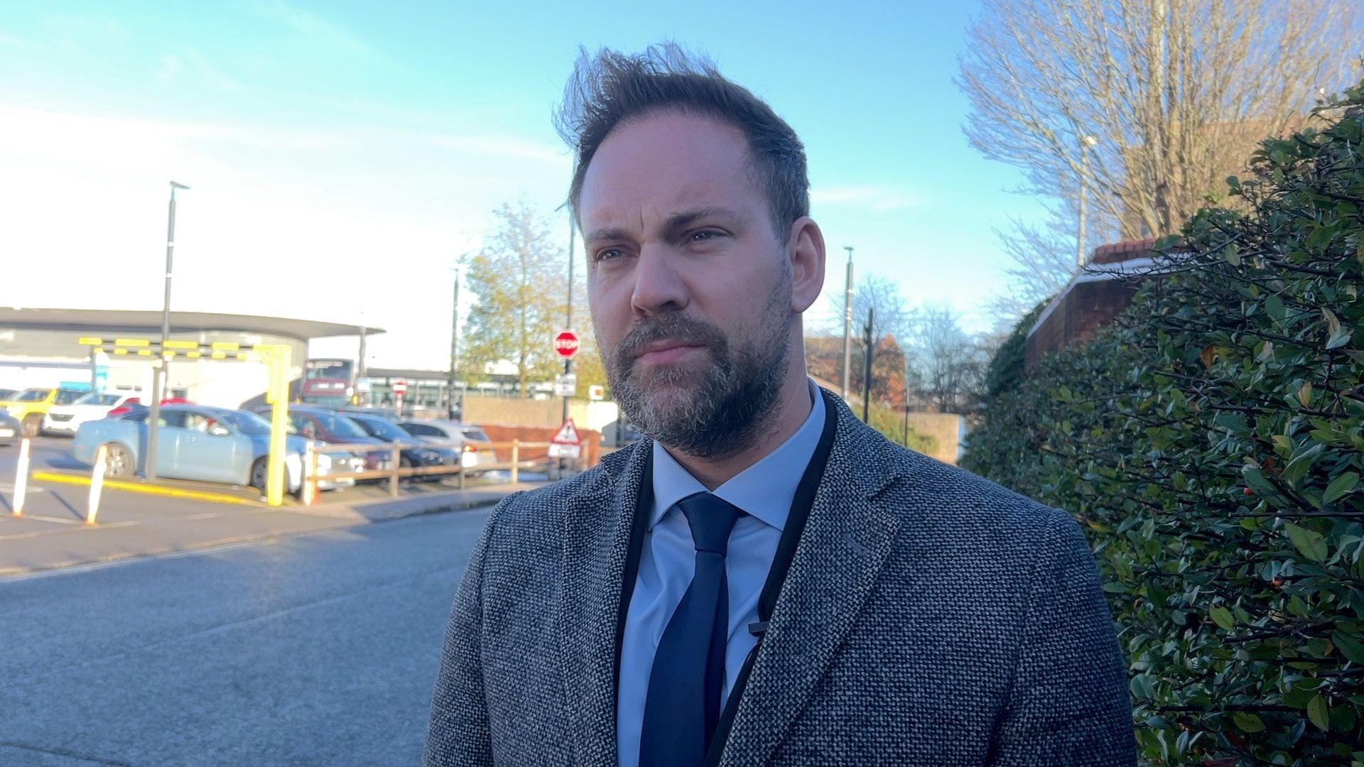 Detective Superintendent Jim Munro wearing a blue tie and grey coat looking into the camera. He is stood in front of a car park with some green bushes to the right of the photo. 