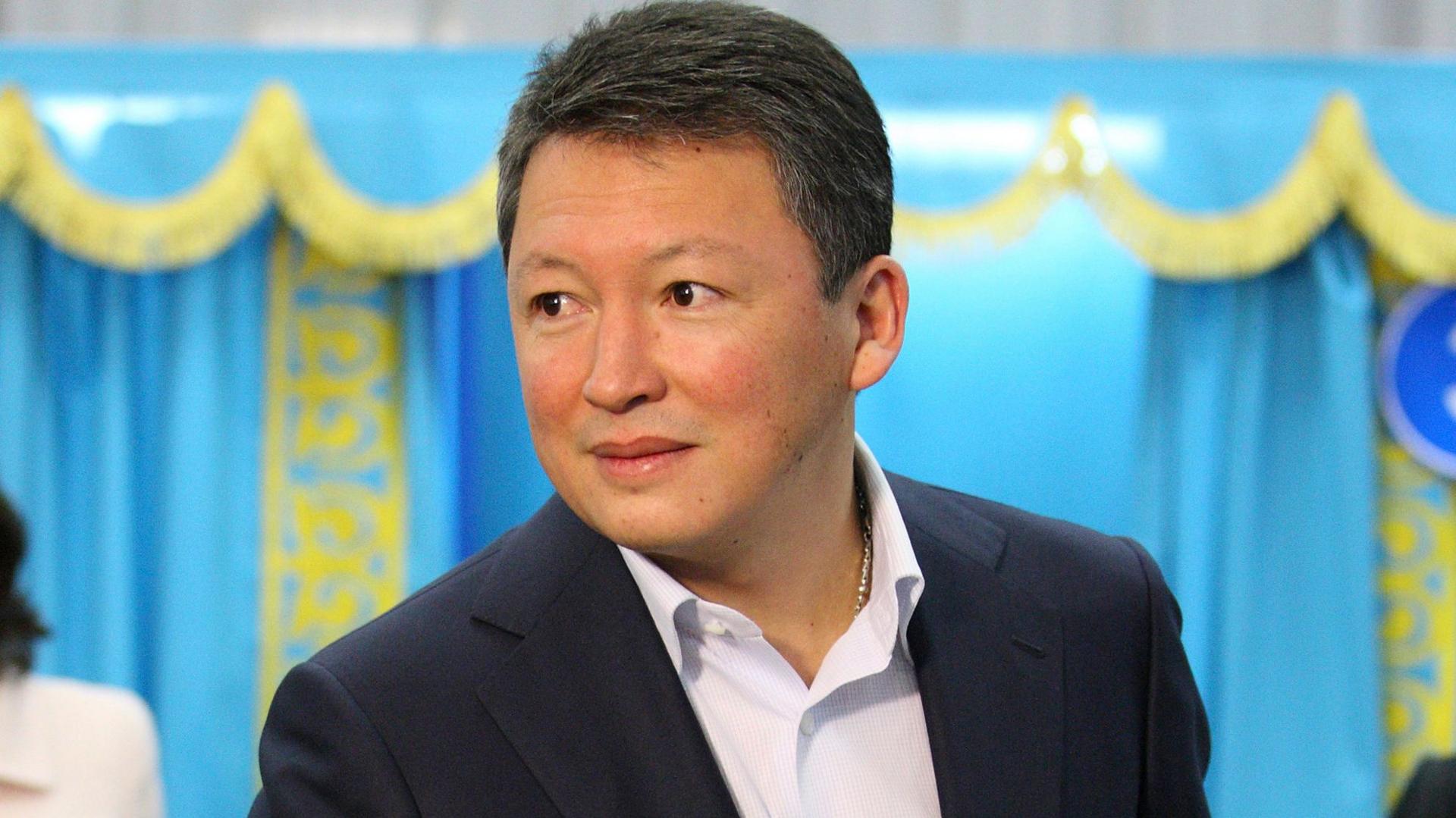 Timur Kulibayev, fotografuar në vitin 2011 duke votuar në zgjedhjet e Kazakistanit. Ai ka veshur një kostum të errët dhe një këmishë të bardhë pa kravatë dhe ka flokë të shkurtër të errët. Pas tij janë perde në ngjyrë blu me zbukurime të arta - ngjyrat e flamurit të Kazakistanit.