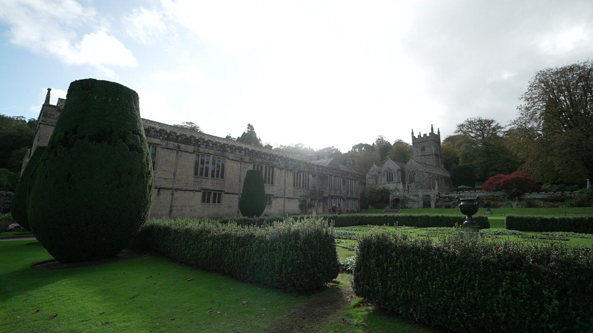 Lanhydrock seen with bushes surrounding the building, there is a castle-type building on the right with turrets, there is a large hedge on the right which has been shaped.