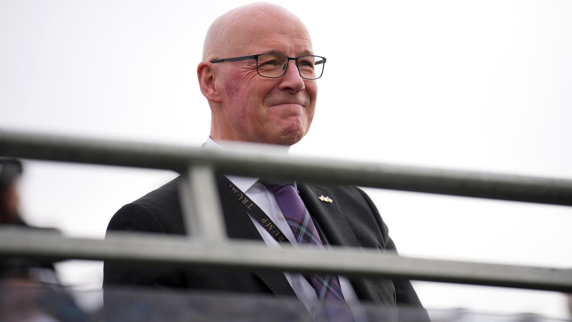 John Swinney, who wears a dark suit with a tartan tie, arrives on a golf course. He is bald, wears glasses and is smiling in the picture. Swinney is sitting down.