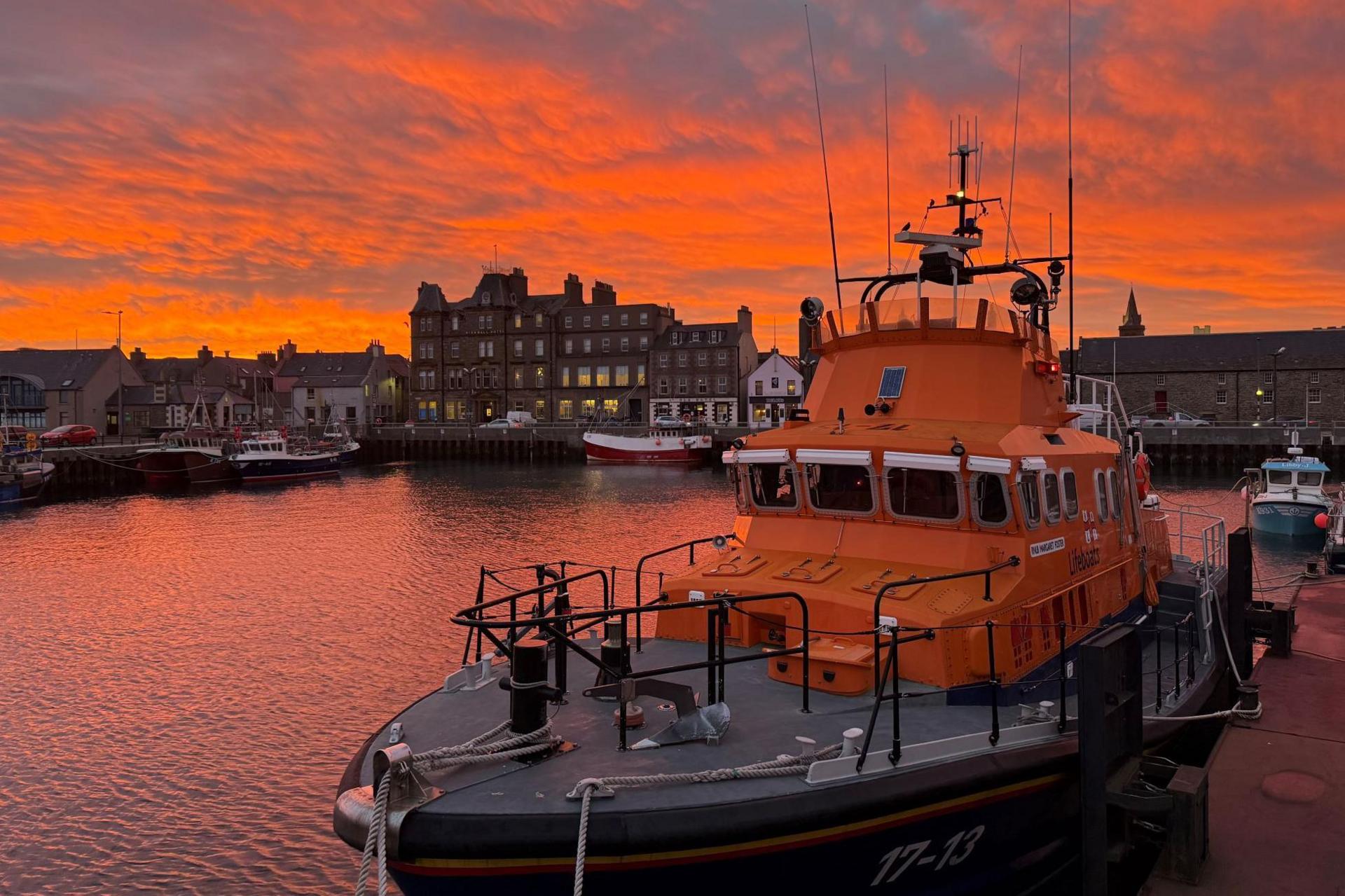 The lifeboat is moored in the harbour under a bright orange sky that matches the colour of the search and rescue boat. There are buildings with lights on in the background.