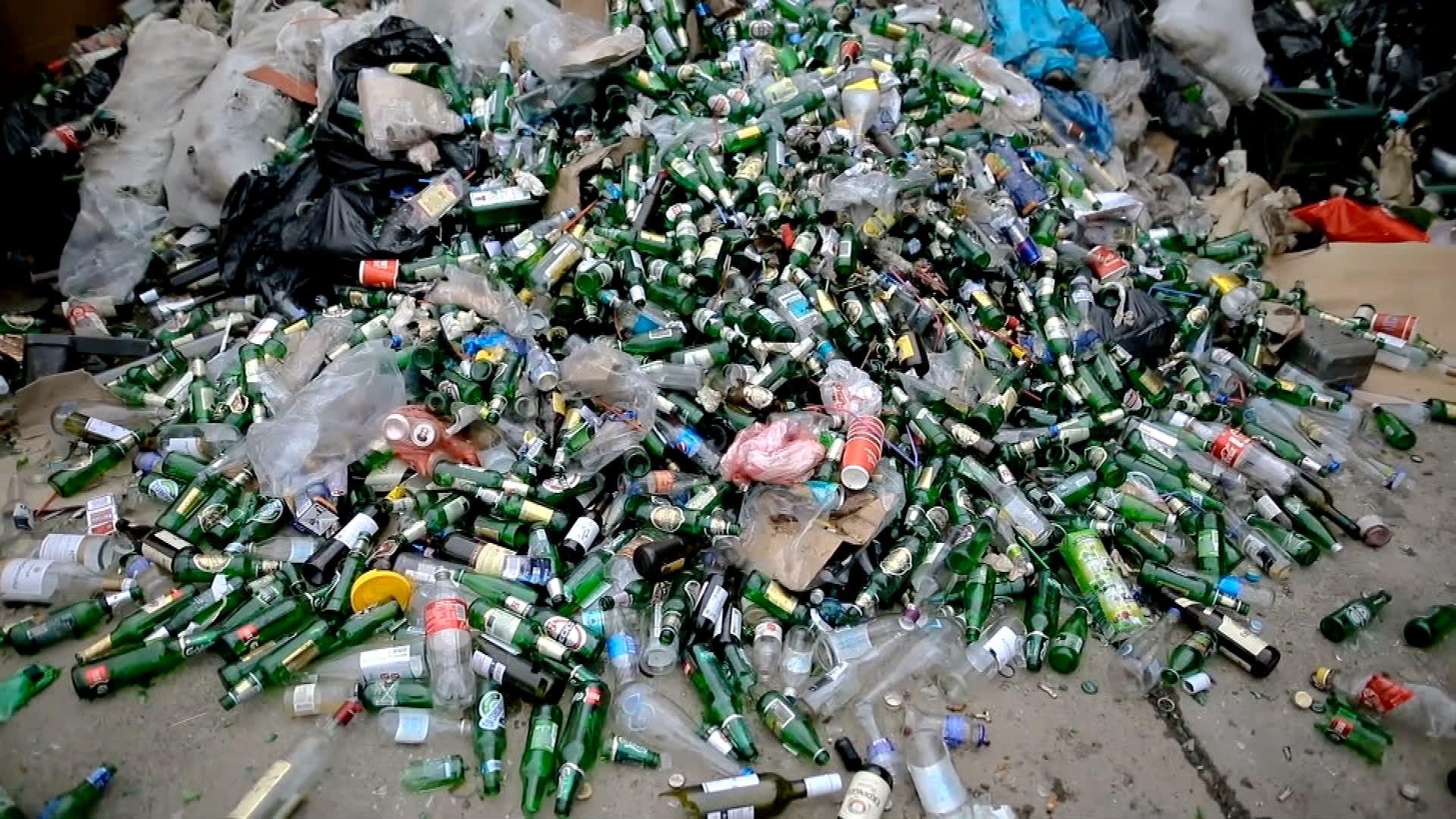 A large pile of glass bottles and a few plastic bags lie in a heap on a concrete floor