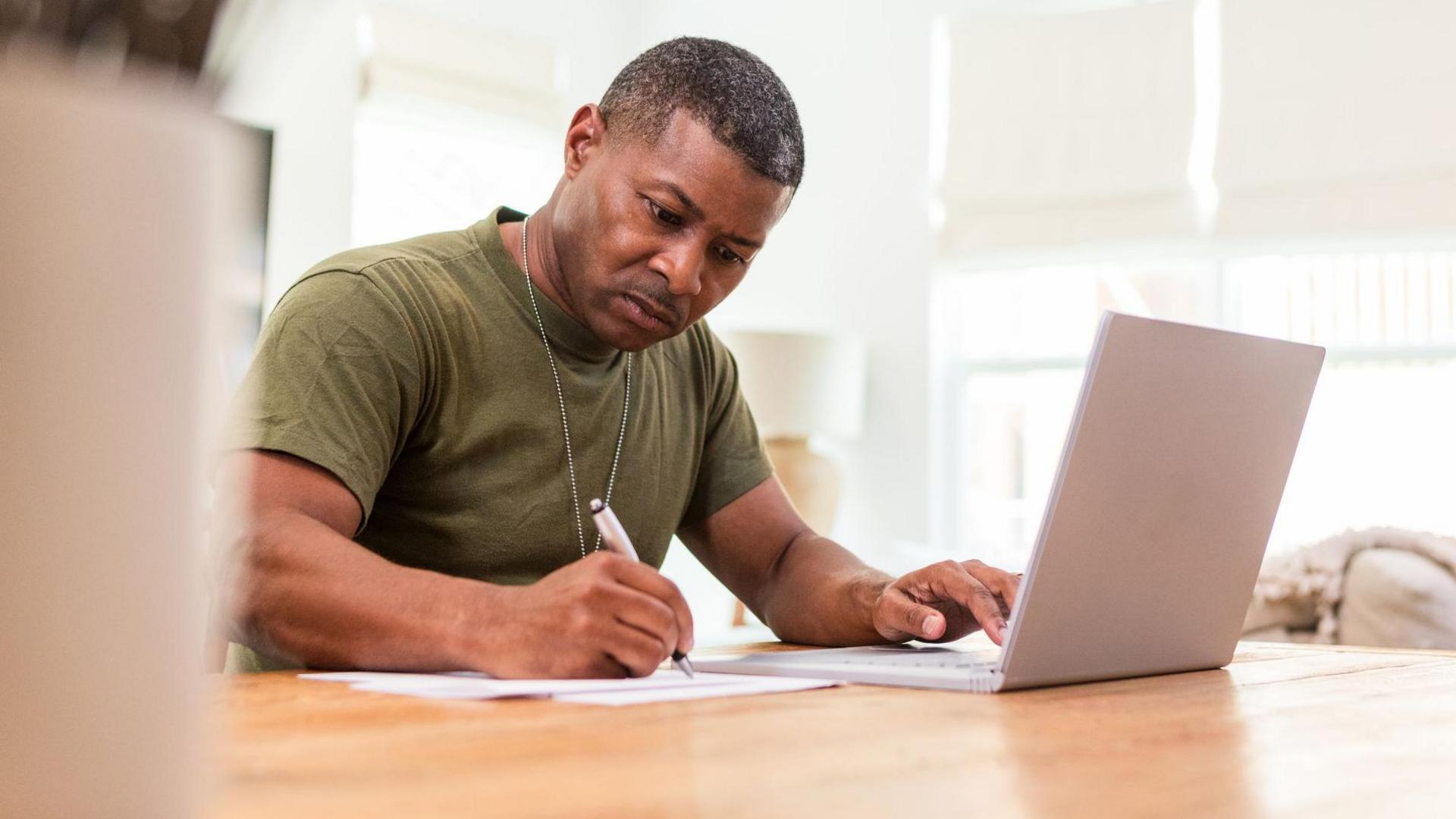 A middle-aged veteran works at a laptop while writing on a piece of paper at a table. He wears a green t-shirt and a dog tag. 