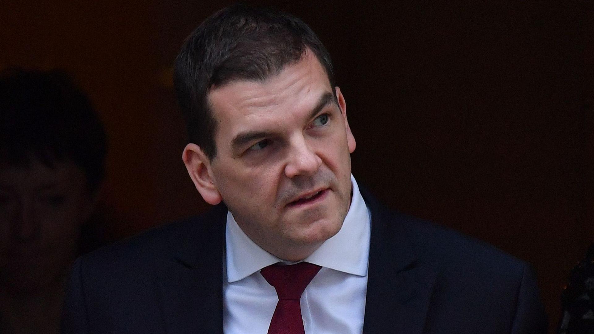 Sir Olly Robbins is wearing a dark-coloured suit, white shirt and a red tie. He has short, black hair.