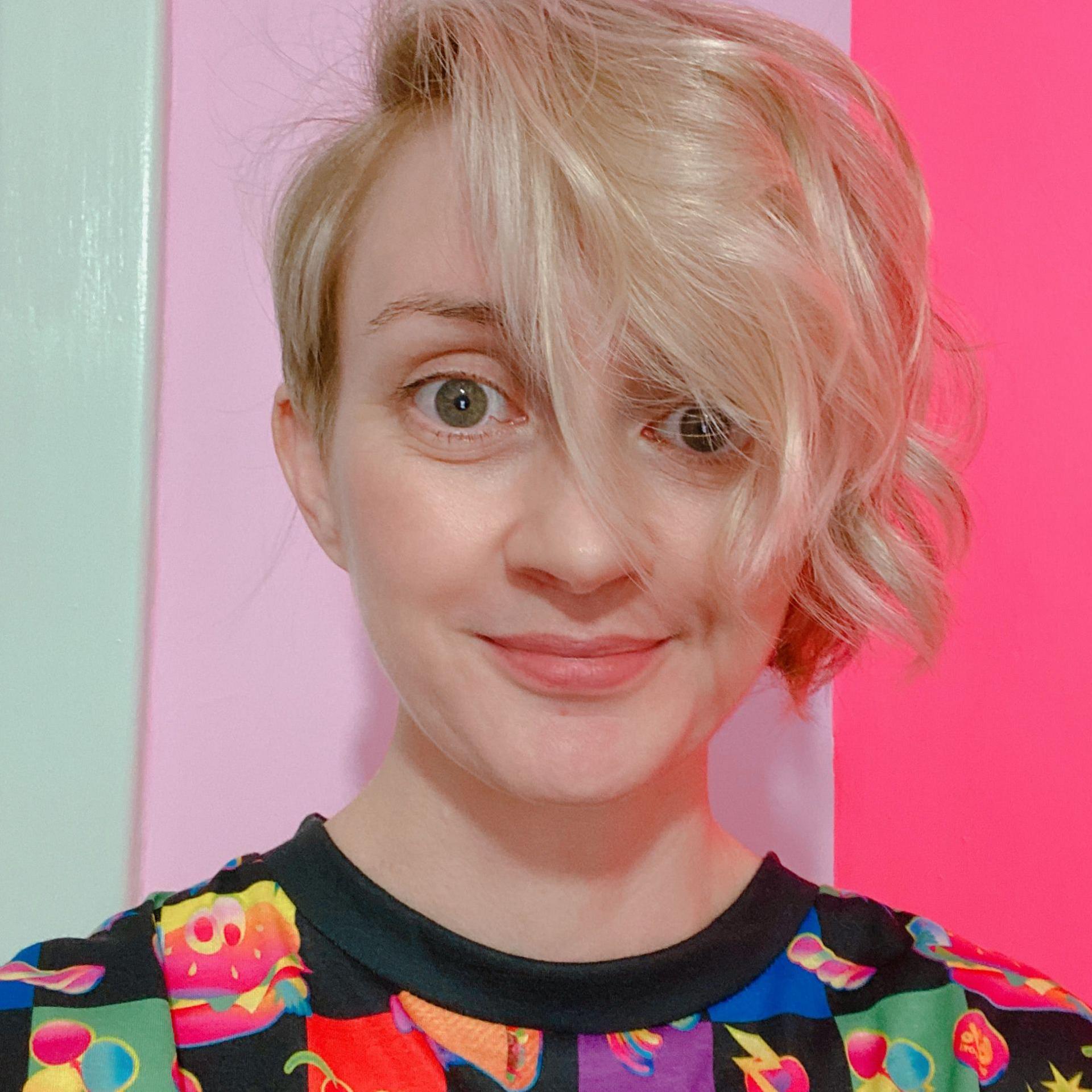 A woman with blond hair and wearing a brightly multi-coloured top. She is standing in front of a pastel backdrop of purples and pinks.