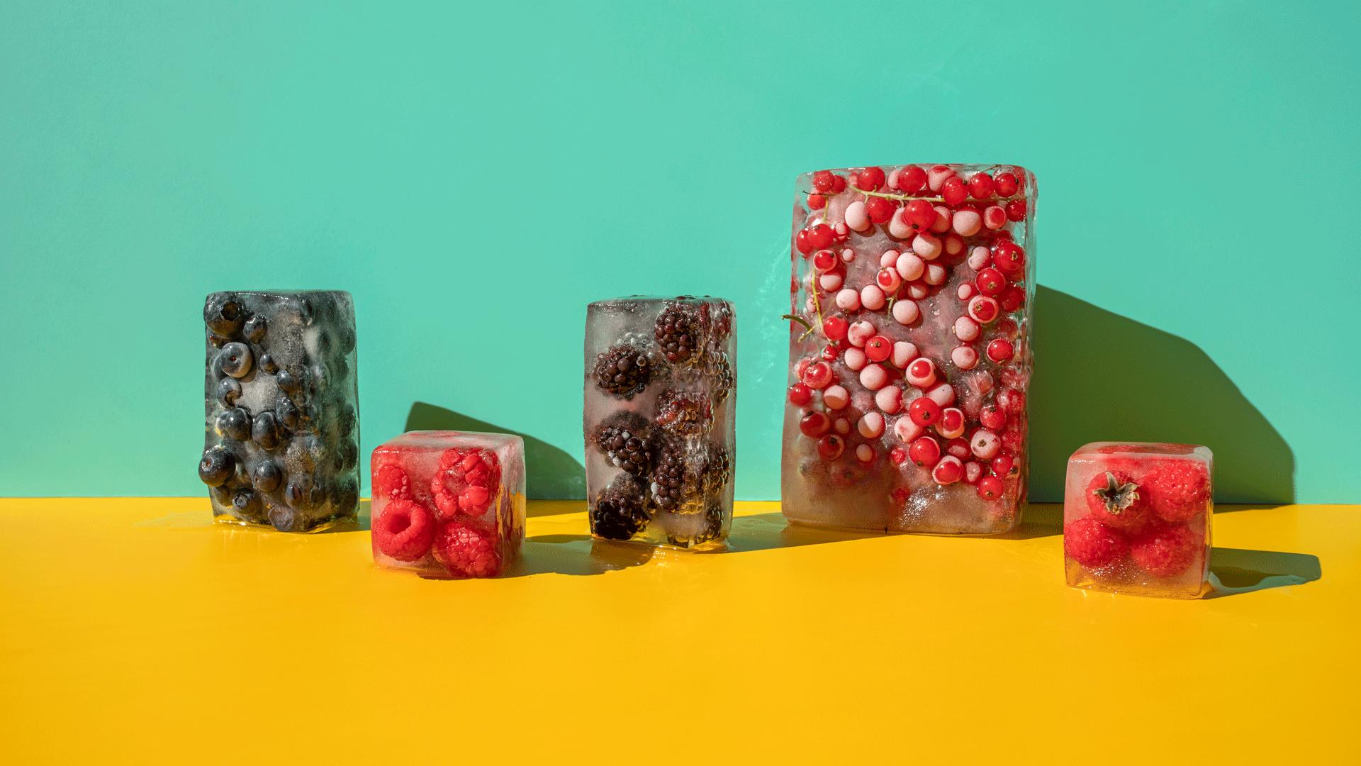 Five blocks of frozen fruit in ice, l-r: medium sized block of blueberries, small sized block of raspberries, medium sized block of blackberries, large sized block of red currents and small sized block of strawberries. They sit on a bright yellow surface with a turquoise background. 