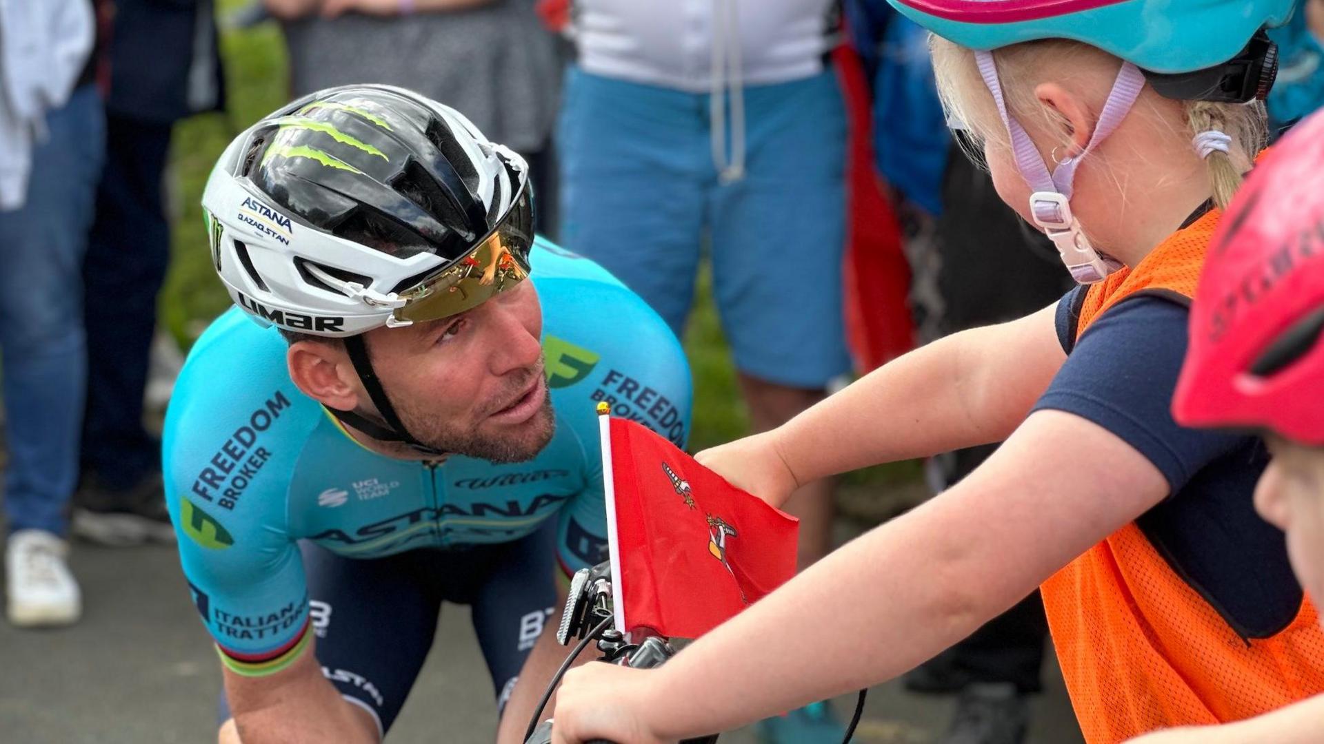 Sir Mark Cavendish in his blue 35 stage victory jersey and helmet, crouching down speaking to a little girl on a bike holding a small Manx flag.