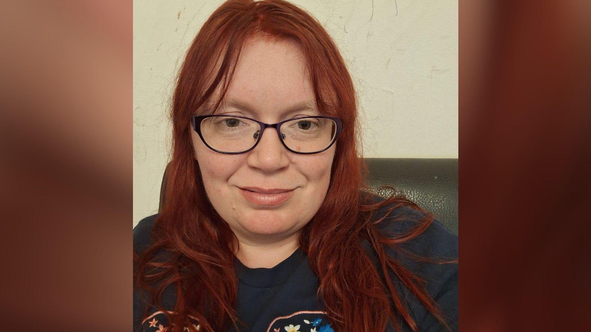 Deborah wearing a navy shirt - black rimmed glasses smiling for her selfie picture. She has red hair and is sitting indoors.