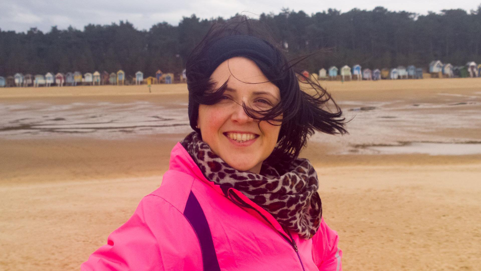 Dr Rachel Gibson posing for a photo at Wells beach in Norfolk. She is smiling at the camera and wearing a bright pink coat, leopard print scarf and has a black fabric hair band on her head. She is standing on the sand with different coloured beach huts seen in the background and with her hair blowing across her face. 