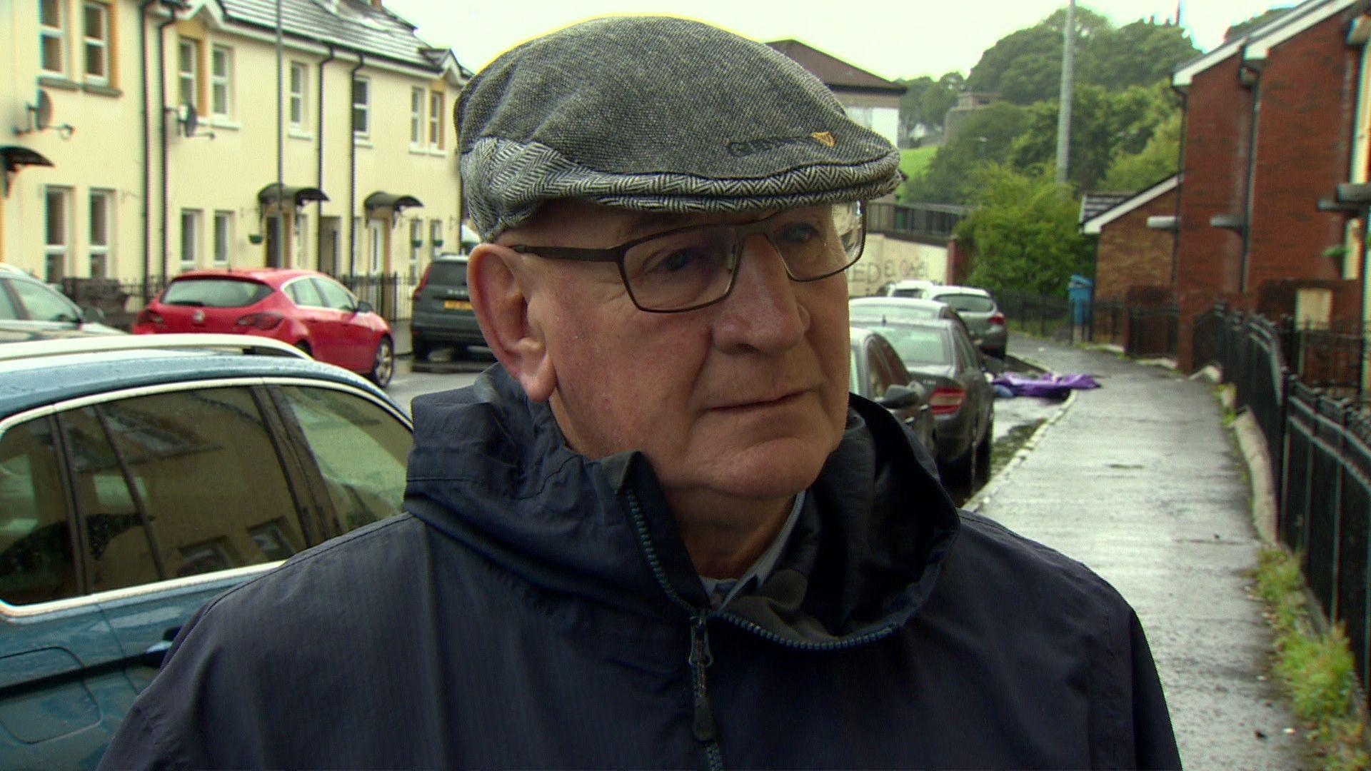 Terry Crossan pictured in front of residential buildings, wearing a navy anorak and a grey flat cap