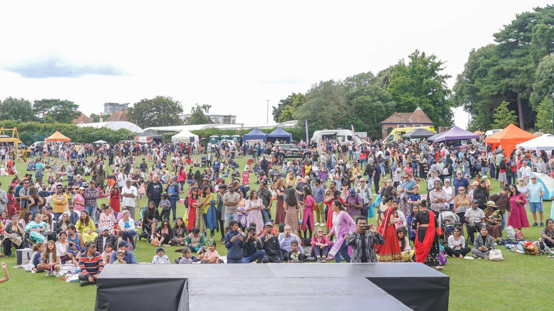 View of an audience from a stage. Hundreds of people sitting and standing in Poole Park.