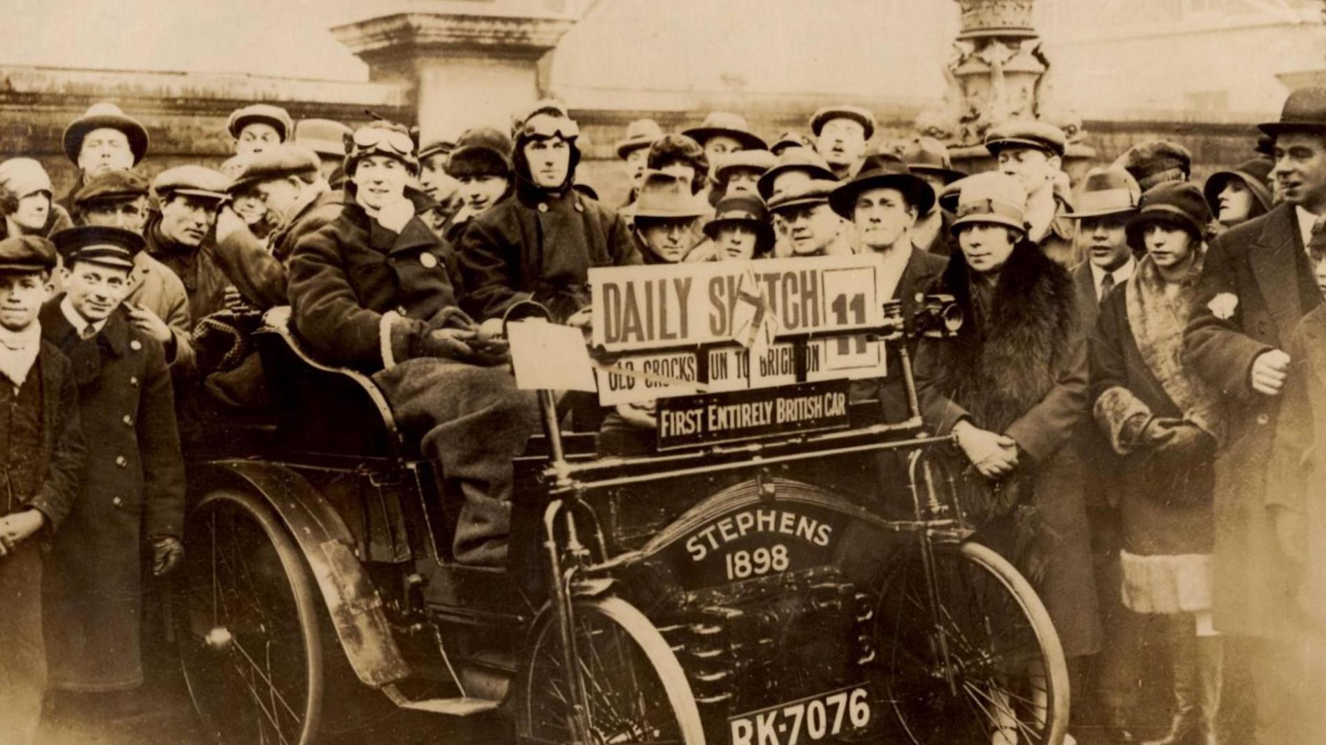 A crowd of people gathered around a Stephens car. All of them are wearing 1920s outfits