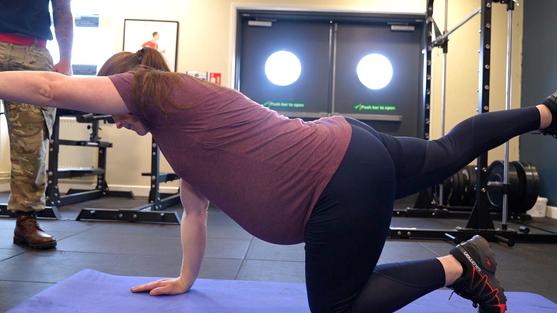 A pregnant woman in purple T-shirt and navy leggings doing floor exercises.