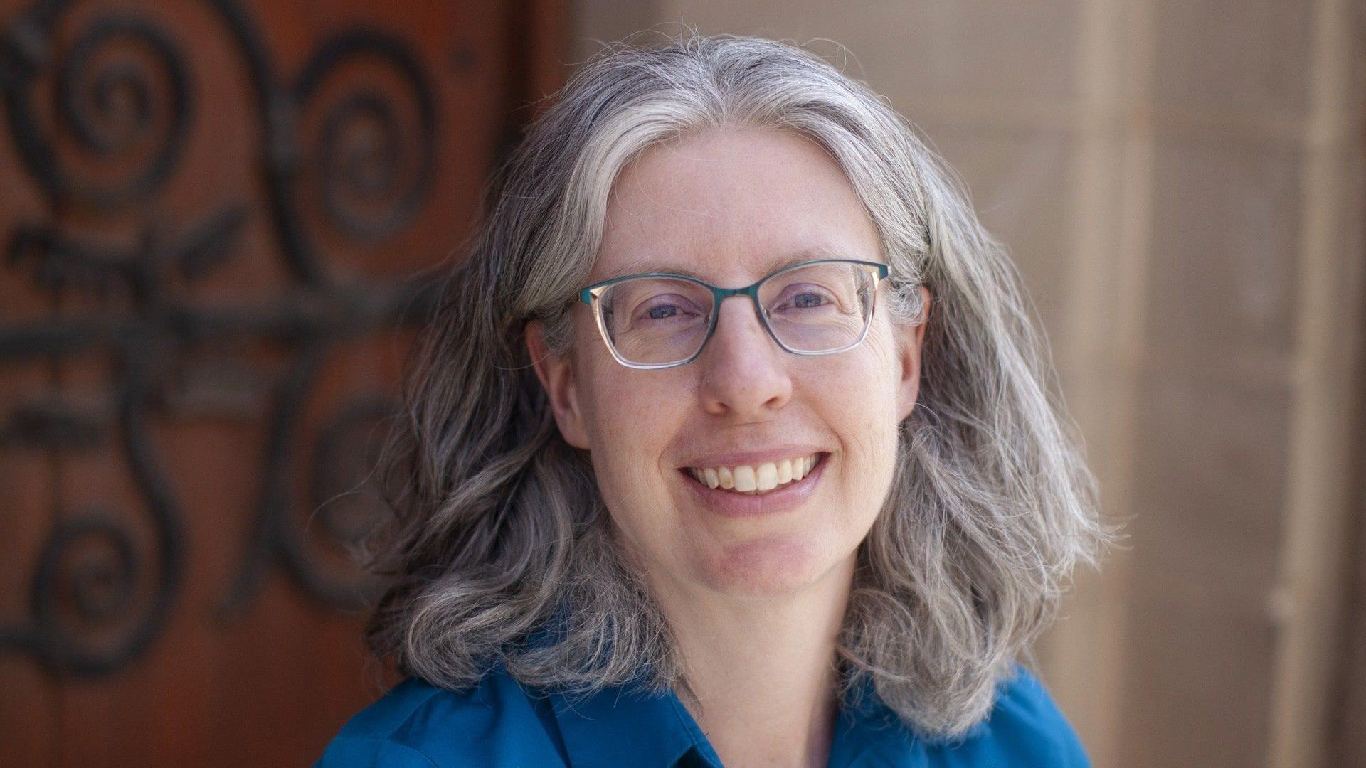 A woman with grey hair and glasses smiles in front of a grand wooden door