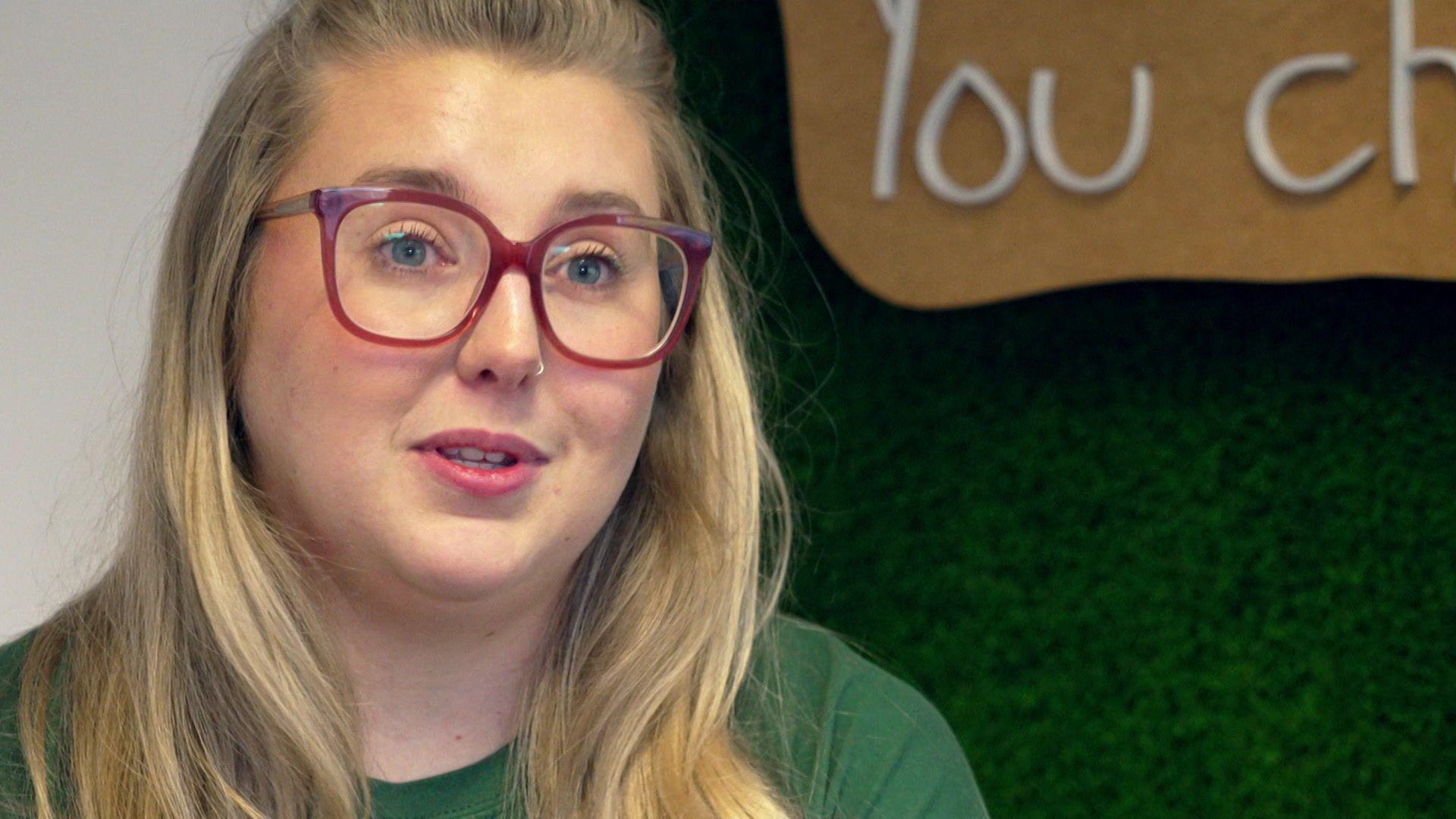Lucy has long blonde hair and blue eyes. She is wearing redframed glasses and a green t-shirt.