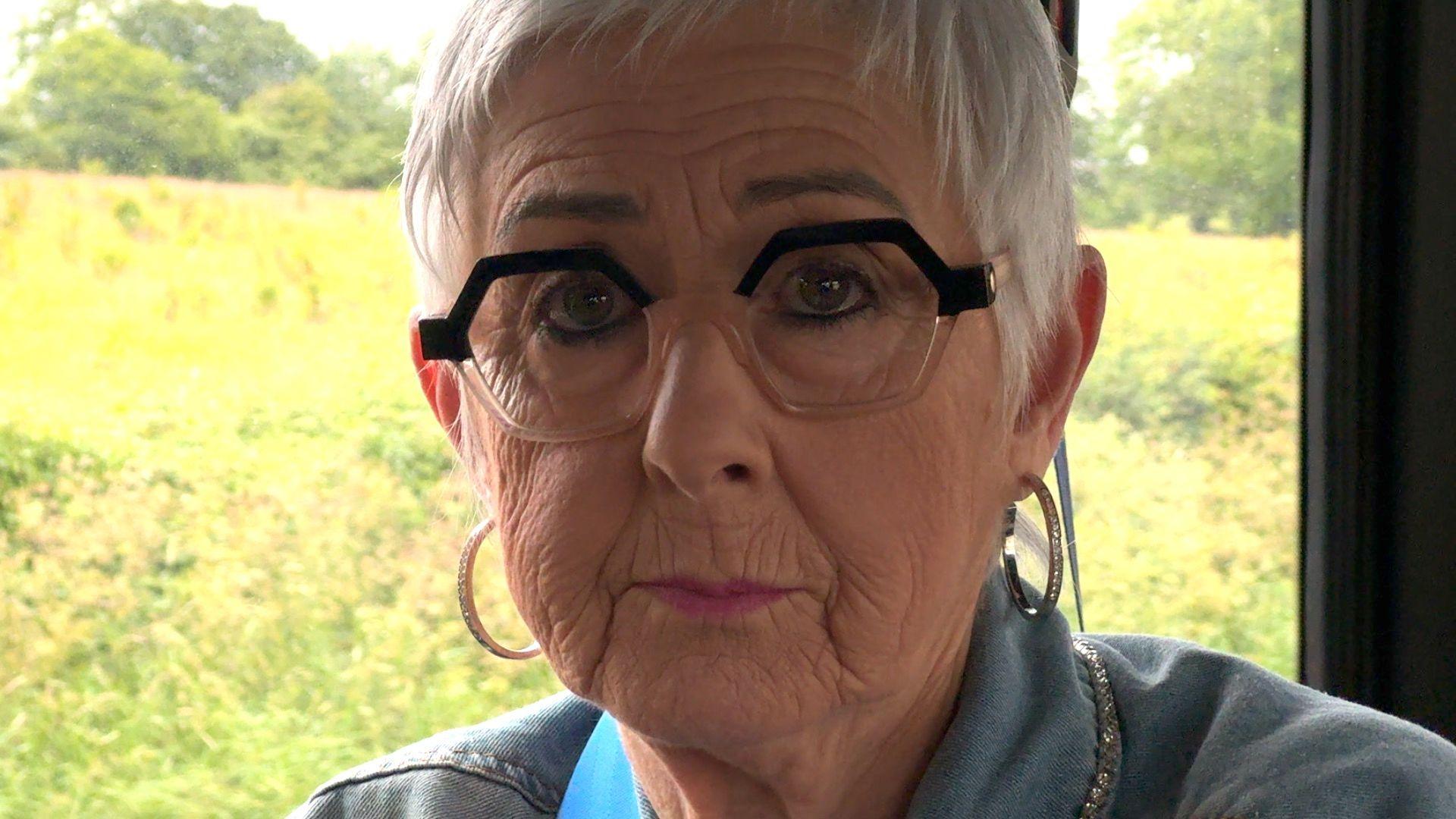 Patricia Davis is sitting against a backdrop of green fields. She has short grey hair and she is wearing distinctive hexagonal-shaped glasses with a black rim 