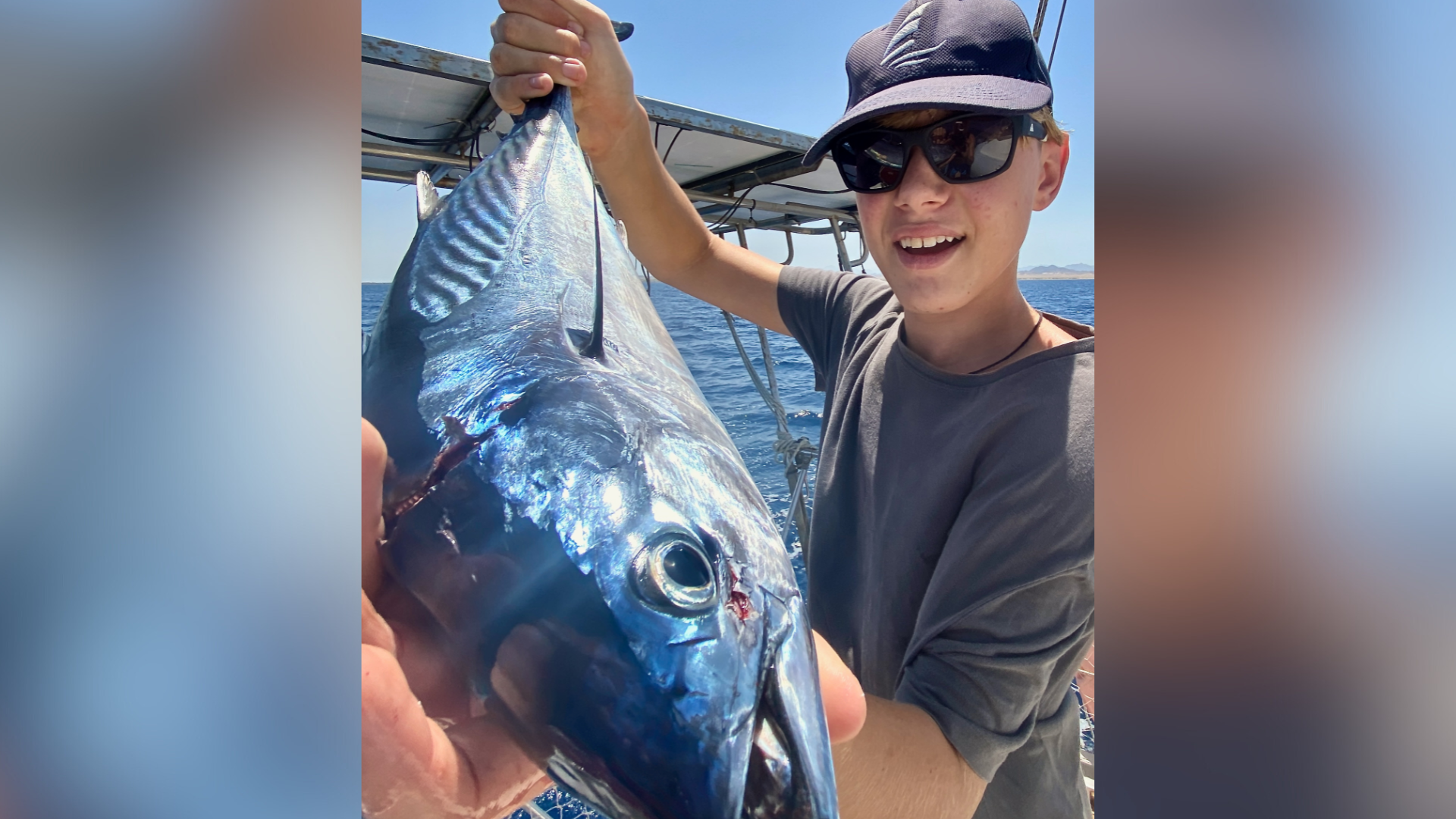 A boy wearing a cap and sunglasses. He is standing on a boat and holding a fish he had caught.