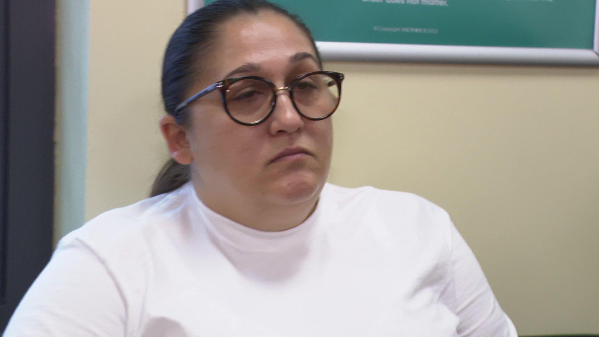 A woman with dark hair tied back and dark round-framed glasses is wearing a white long-sleeved top and sitting in front of a yellow wall.