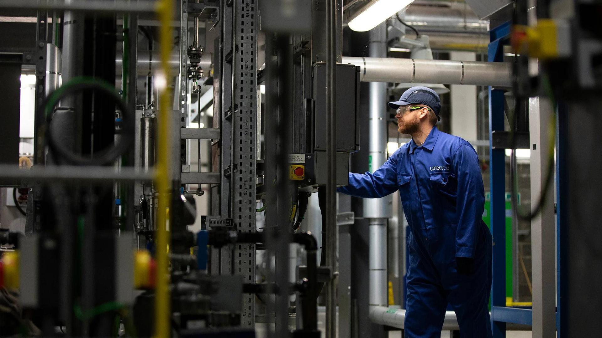 A worker wearing a hat and navy overalls looking at machinery in a factory