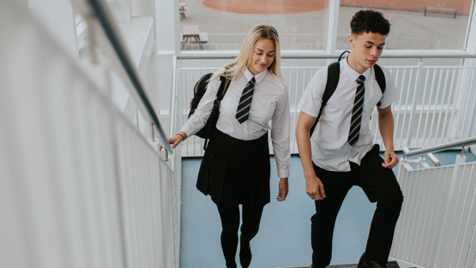 A girl (left) and a boy (right) walking up indoor school steps wearing school shirt and tie.