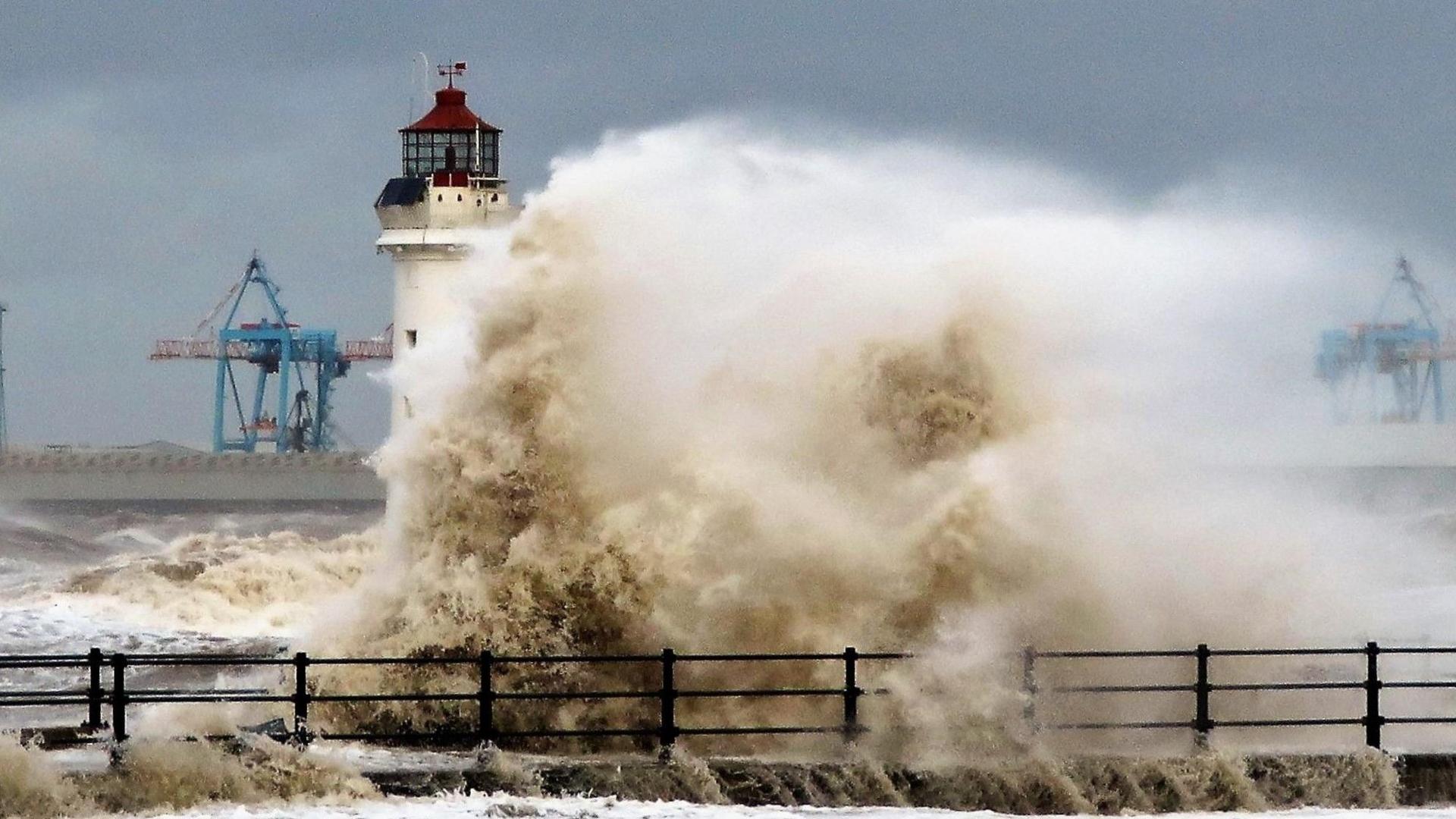 Waves surge against a white lighthouse in Merseyside, engulfing its base in spray as a storm batters the coastline. Industrial cranes from a nearby port are visible in the background.